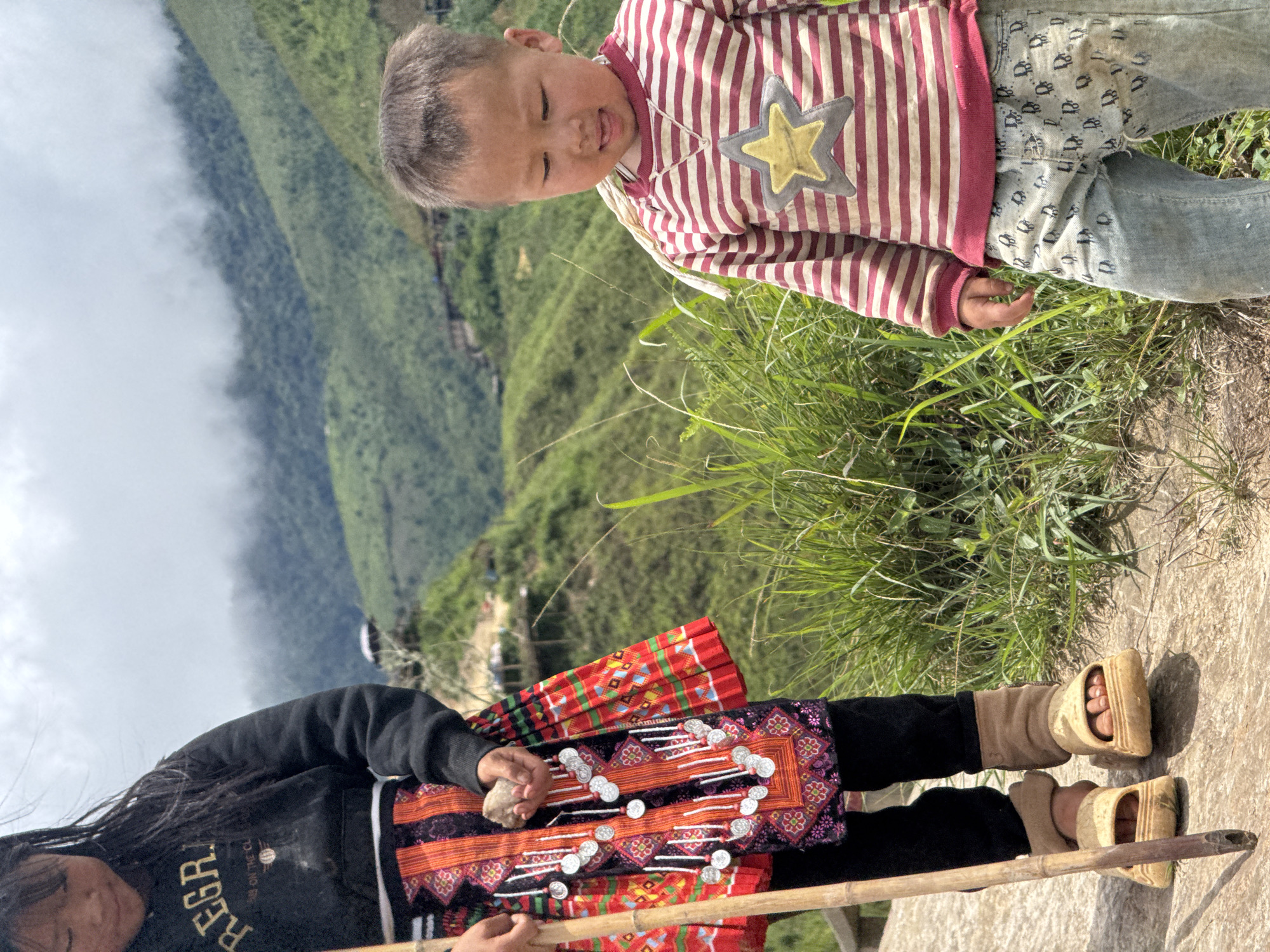 In Bắc Yên, Vietnam, a young child with dark hair and a black hoodie stands on a path, holding a bamboo stick. They wear a colorful, patterned skirt adorned with silver decorative elements. To their right, a younger child in a striped long-sleeved shirt and patterned jeans looks down. The background features lush green mountains shrouded in mist, with scattered buildings visible on the slopes.