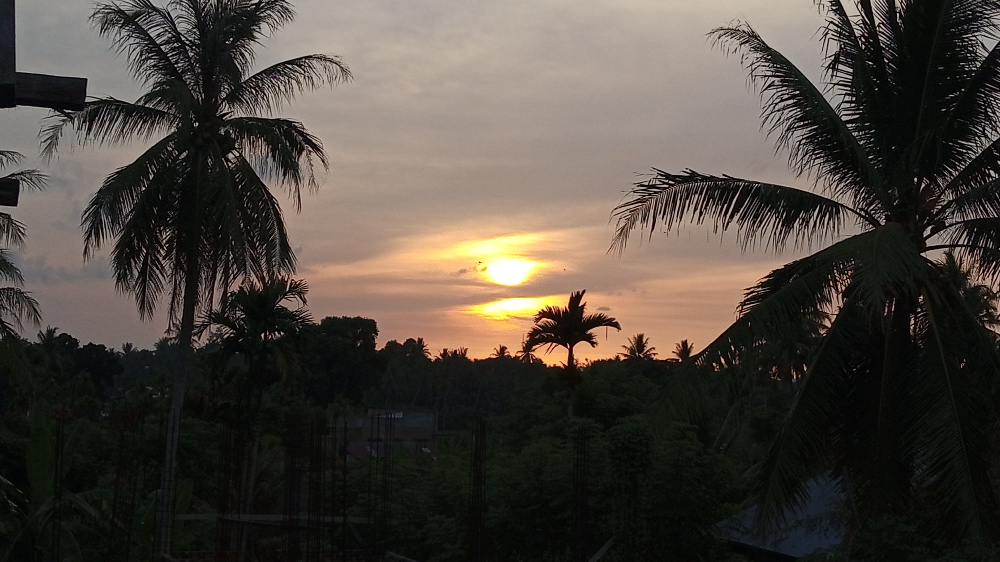 The image depicts a sunset scene in Buketteukuh, Indonesia, with the sky filled with soft, warm hues of orange and yellow. The sun, a bright, luminous orb, is partially obscured by clouds, casting a golden glow across the sky. In the foreground, silhouetted against the vibrant sky, are the fronds of several palm trees, their leaves creating a delicate pattern. Below the sky, a dense canopy of green foliage stretches across the landscape, indicative of a tropical environment. The trees are lush and varied, suggesting a natural, possibly forested area. In the middle ground, hints of structures, possibly houses or buildings, can be discerned amidst the greenery, suggesting a rural or semi-urban setting within Buketteukuh. The overall impression is one of tranquility and natural beauty, capturing the serene end of a day.
