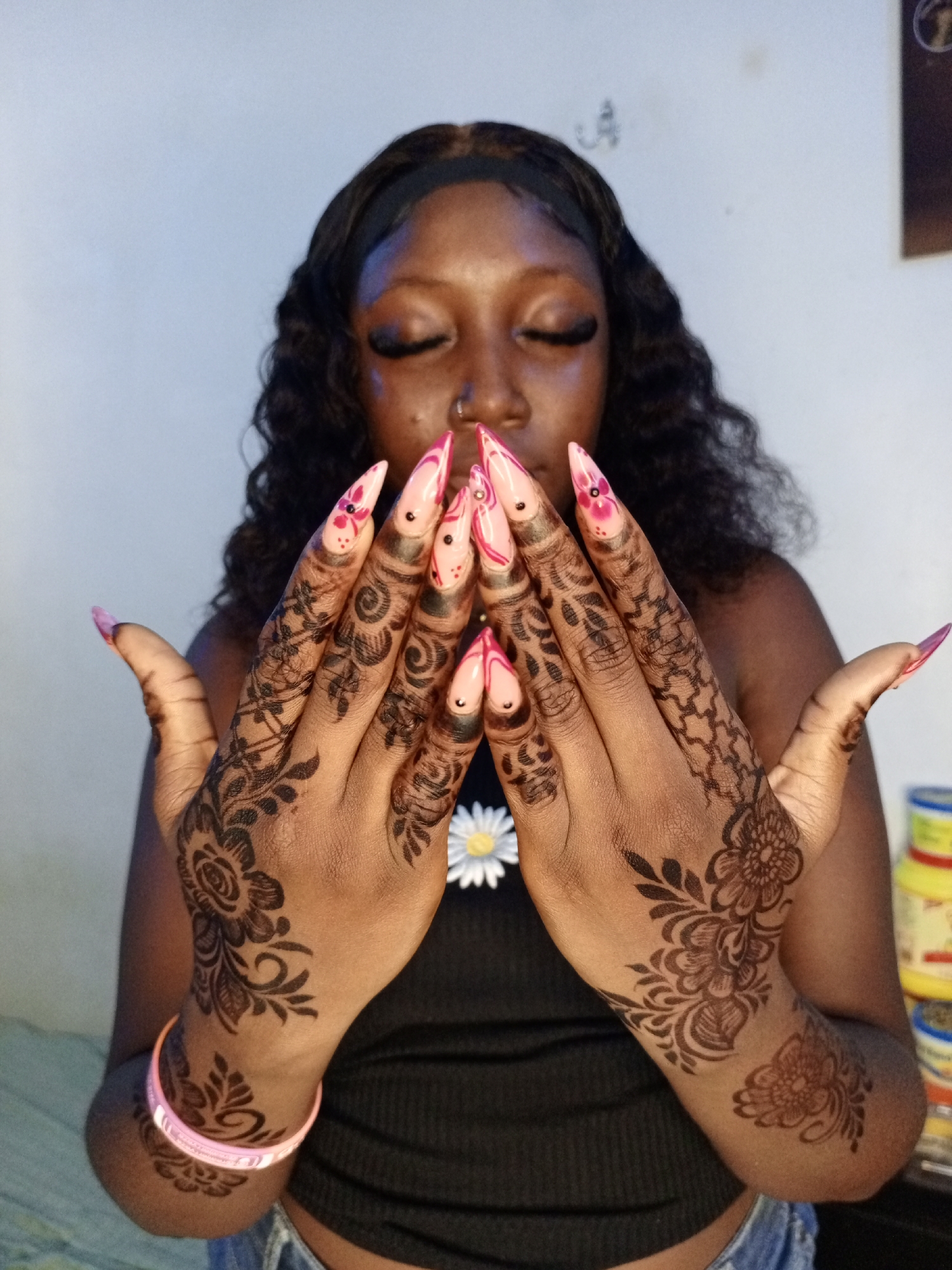 A young Black woman with her eyes closed is displaying both hands toward the camera. Her fingernails are long, almond-shaped, and painted in light pink and dark pink floral designs. Henna tattoos in a traditional pattern cover the back of her hands and forearms. She is wearing a black tank top with a small white flower on the left breast and a light pink bracelet on her left wrist. Her hair is long, black, and wavy. The setting is an indoor room of unknown location in Unknown, Unknown.
