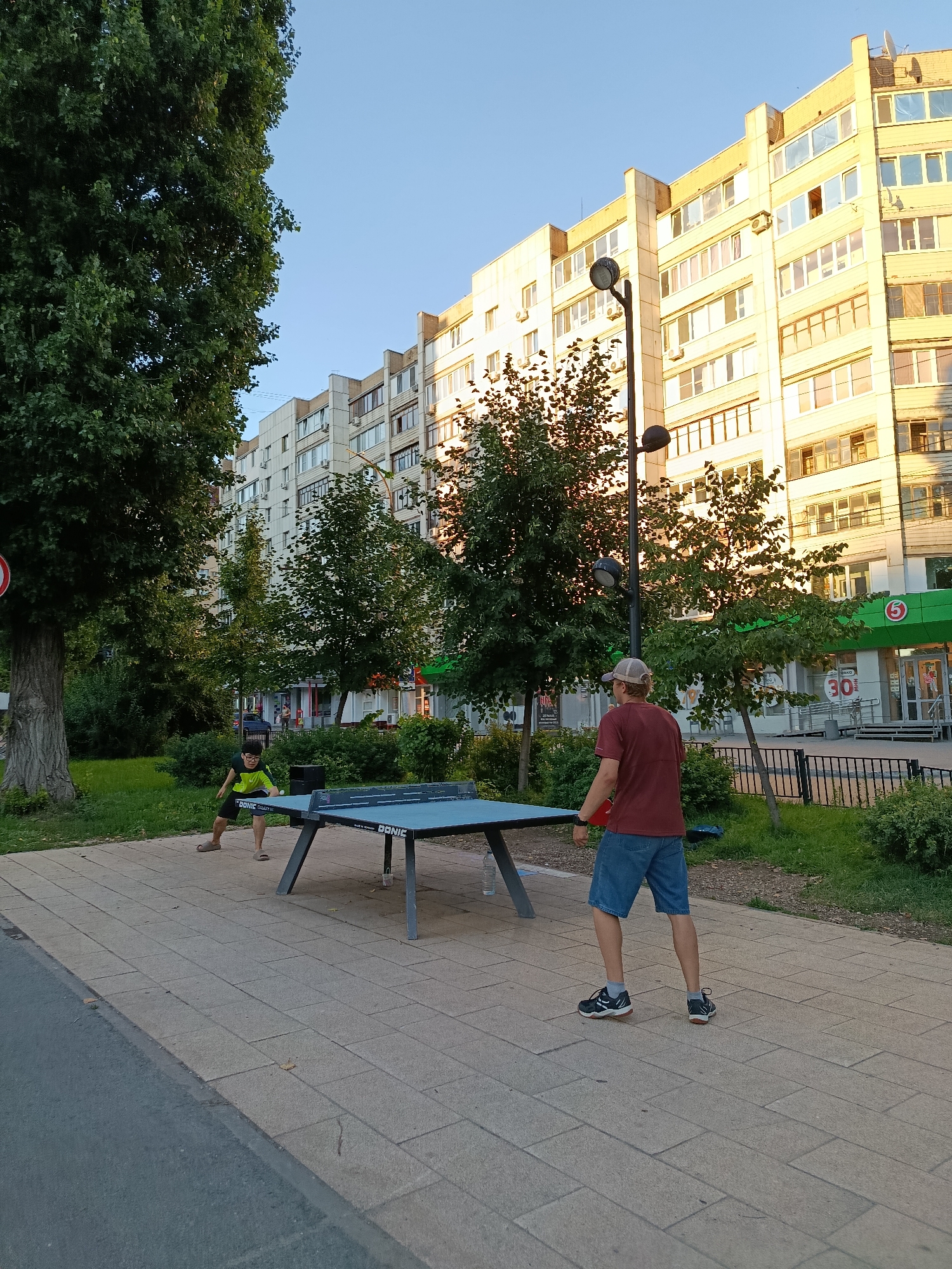 Two individuals are playing table tennis on a public table set up on a paved area adjacent to a residential building. The person closer to the camera, wearing a maroon t-shirt and denim shorts, stands with their back to the viewer, holding a red paddle. The other person, a child wearing a green shirt and dark shorts, is on the opposite side of the table, actively engaged in the game. The table tennis table is blue with "BONIC" branding. Surrounding the playing area are trees, green grass, and a metal fence. In the background, a multi-story residential building with many windows dominates the skyline, with a supermarket visible at its base. The scene appears to be set in the late afternoon or early evening, as indicated by the warm lighting and long shadows. The location is confirmed to be Saratov, Russia, based on the architecture and signage.