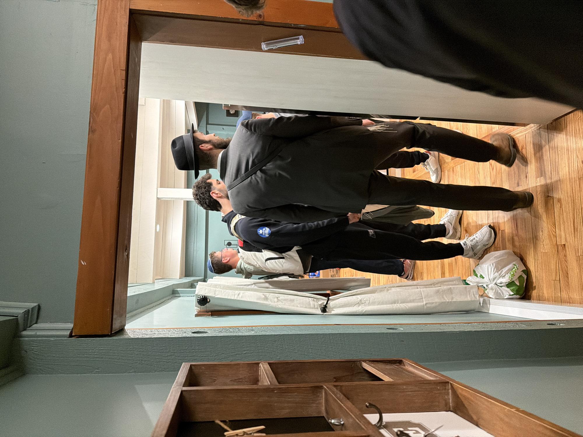 A group of people, including a man in a suit and hat, stand inside a room with light blue walls and wooden flooring. The room is in Rock Hill, United States of America. A partially visible wooden box is in the foreground. A set of blinds, rolled up, is in the middle of the room. A plastic bag rests on the floor near the blinds.