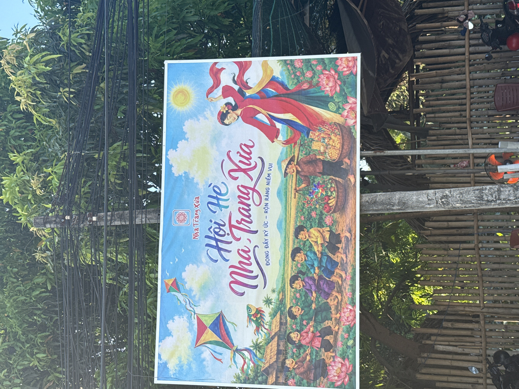 A billboard is displayed outdoors, depicting a vibrant scene with a blue sky, fluffy clouds, and a bright sun. The billboard features a traditional Vietnamese village with thatched-roof houses, children playing tug-of-war, and a woman in a red dress dancing with flowing red scarves. Kites are flying in the sky, and lotus flowers are blooming in the foreground. The text on the billboard is in Vietnamese and appears to advertise a cultural event or attraction. The billboard is mounted on utility poles, with lush green trees and tangled wires overhead. The surrounding environment includes a bamboo fence, a motorbike, and a rustic thatched-roof structure. The overall impression is a celebration of Vietnamese culture and a depiction of a joyful, traditional setting. This scene is located in Nam Nha Trang Ward, Vietnam, as evidenced by the billboard's text and imagery.