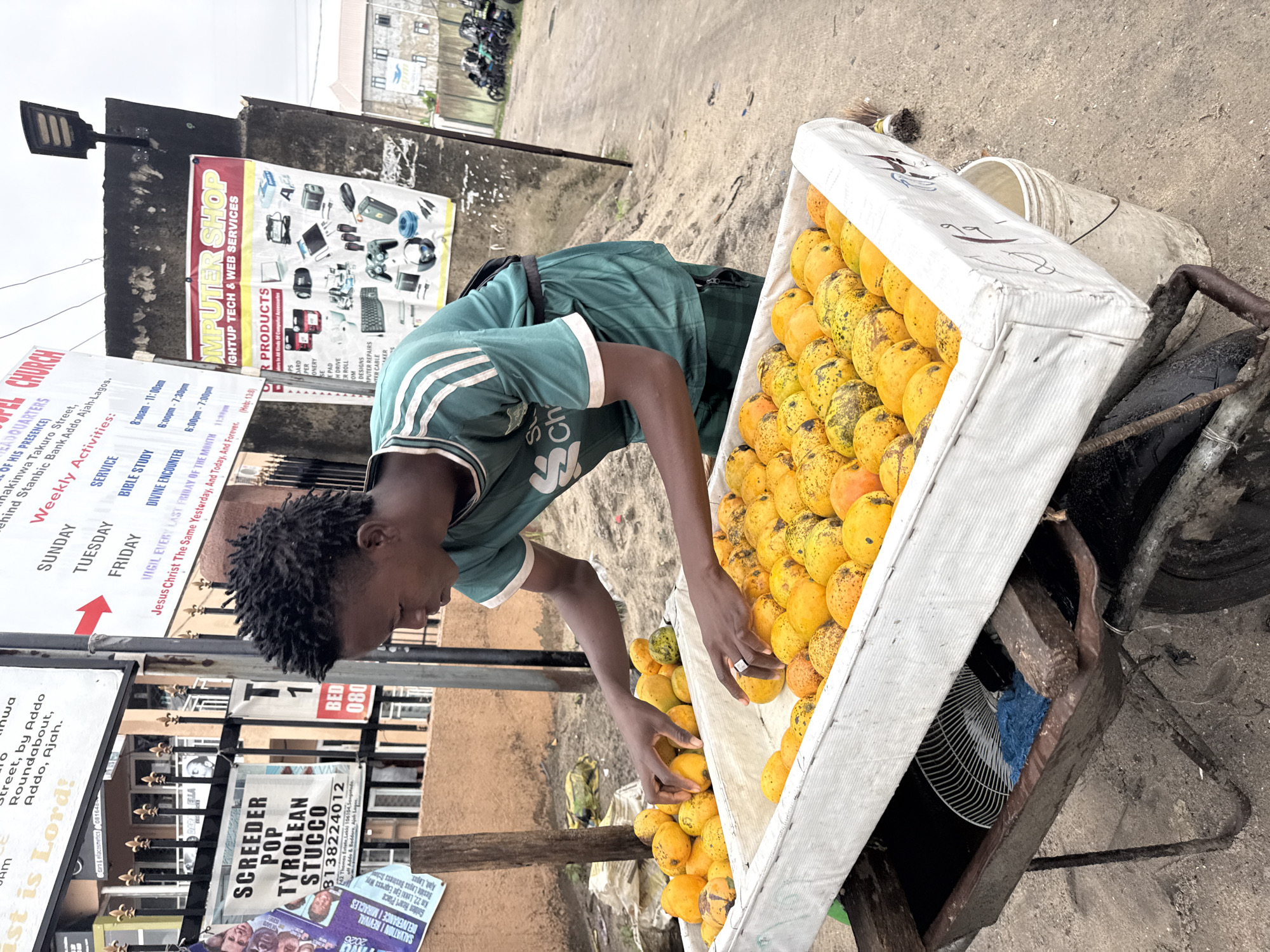 A young man with dark skin and dreadlocks wearing a green and white shirt is selecting mangoes from a cart on a street in Ado, Nigeria. The cart is overflowing with yellow mangoes. A sign for "Screeder Pop Tyrolean Stucco" is visible, with a phone number. A computer shop advertisement and church sign are in the background. The environment appears to be a typical urban street.