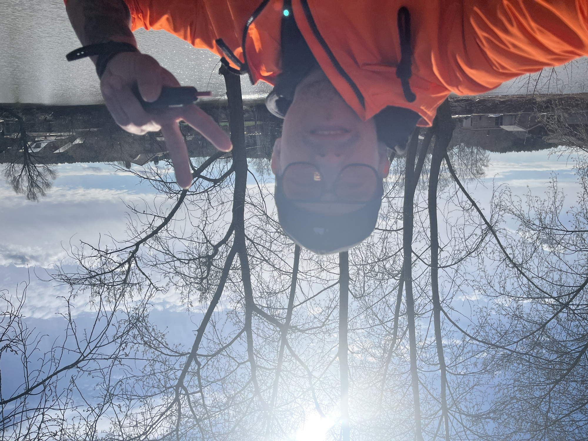 The image displays an inverted selfie taken outdoors, featuring a person in the foreground. The person, appearing male, is wearing a bright orange jacket, dark-rimmed glasses, and a dark cap. They are smiling and making a peace sign with their left hand, on which a dark wristband or watch is visible.
When correctly oriented, the scene places the person against a backdrop of bare tree branches, suggesting a winter or early spring season. Behind the branches and the person, a calm body of water, likely a lake or river, stretches across the middle ground. On the opposite bank, distant trees and some structures or buildings are vaguely visible under a partly cloudy sky. The sky above the far bank is a bright blue with white clouds.
A significant sun flare emanates from the lower right portion of the original (inverted) image, indicating a sunny day. The overall impression is one of a cheerful moment captured in a natural, open environment. There is no visible text in the image. The location context is Chaikovsky, Russia.