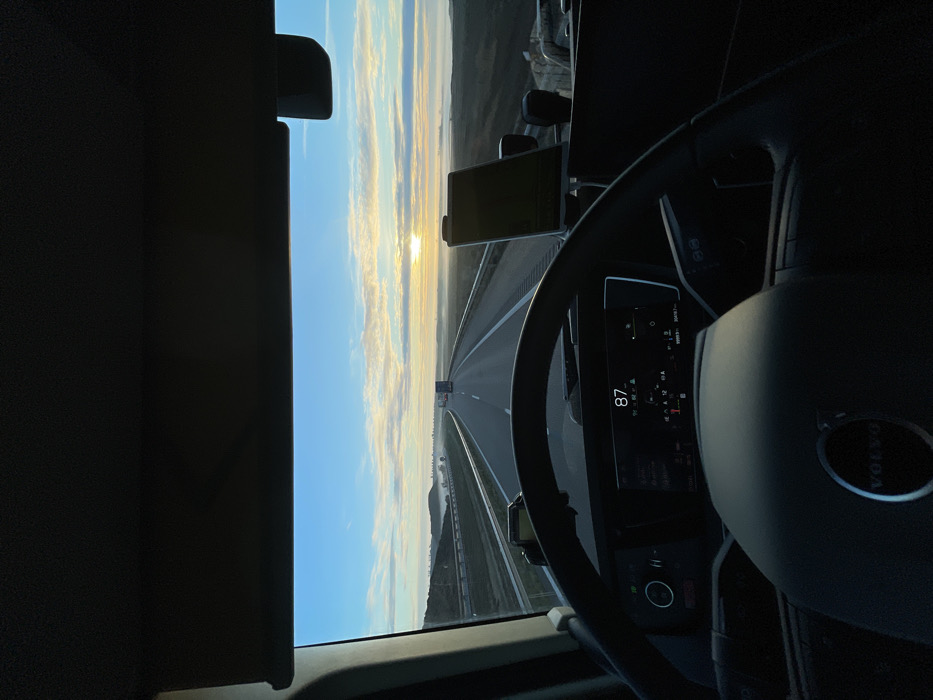 Inside a Volvo truck, the driver's perspective reveals a straight highway stretching towards the horizon under a bright, early morning sky. The rising sun casts a golden glow across the landscape. The dashboard displays a speed of 87, while a tablet sits mounted to the right. The steering wheel partially obscures the digital display. The left side of the frame is dominated by a dark sun visor. Another truck is visible on the road ahead.