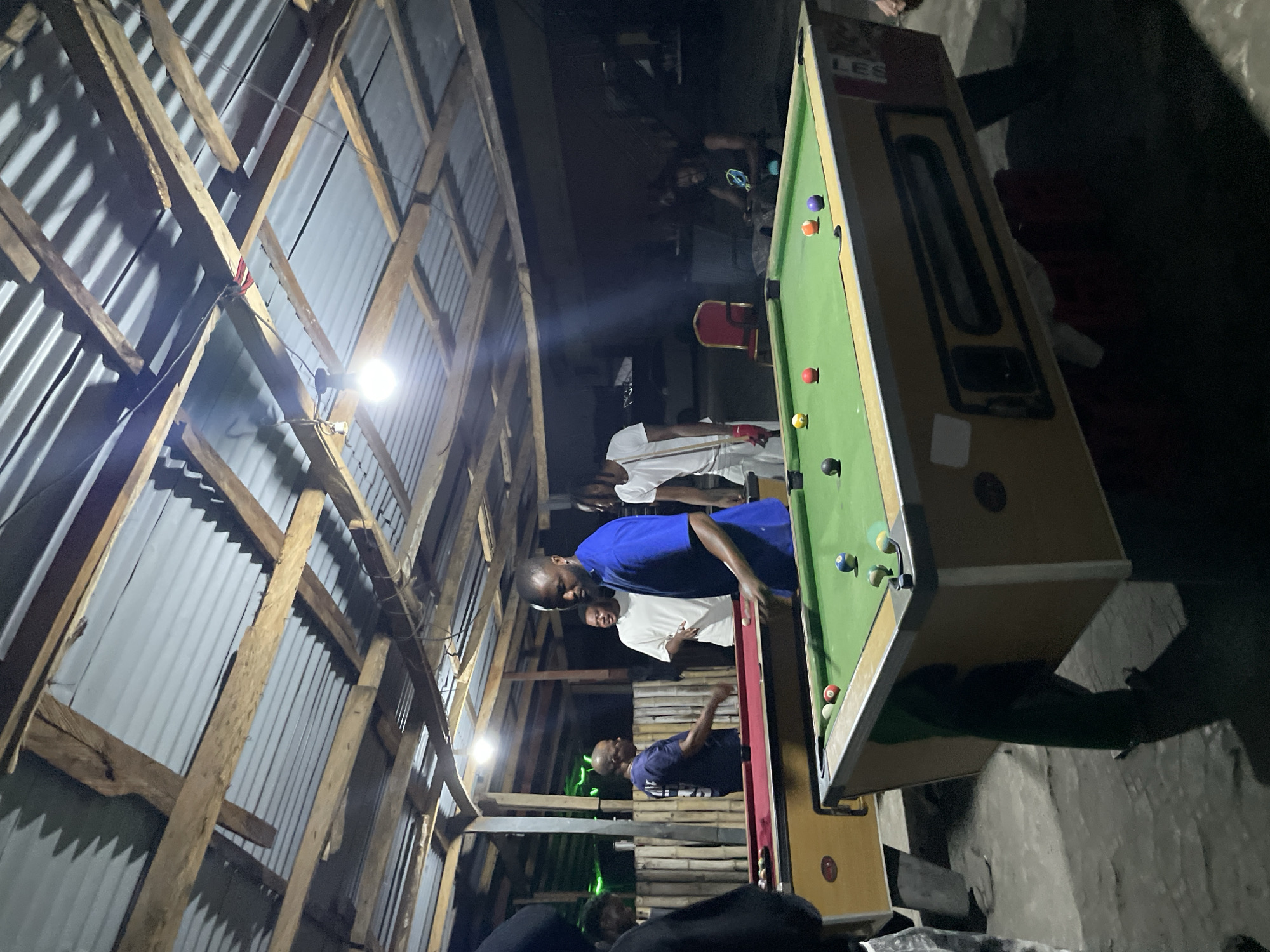 This image, taken in Ado, Nigeria, depicts an indoor scene at what appears to be a casual game room or community space, likely at night given the dim lighting and reliance on artificial light sources.
The setting is characterized by a rustic overhead structure made of wooden beams and corrugated metal sheeting, with bare light bulbs hanging to illuminate the area. A prominent feature is a green-felted pool table, around which several men are gathered.
The central activity is a game of pool. A man in a blue shirt is leaning over the table, intently focused on making a shot. Various colored pool balls, including red, green, yellow, purple, and a black eight-ball, are visible on the table, along with the white cue ball. Other men are positioned around the table, observing the game or waiting for their turn. One individual in a white shirt is also holding a pool cue. In the background, other figures are faintly discernible in the shadows.
Notable details include the informal construction of the building and the simple artificial lighting. A red chair is visible in the background. The text "LES" can be seen on the side of the pool table. The overall mood suggests a relaxed, social gathering centered around the game of billiards.