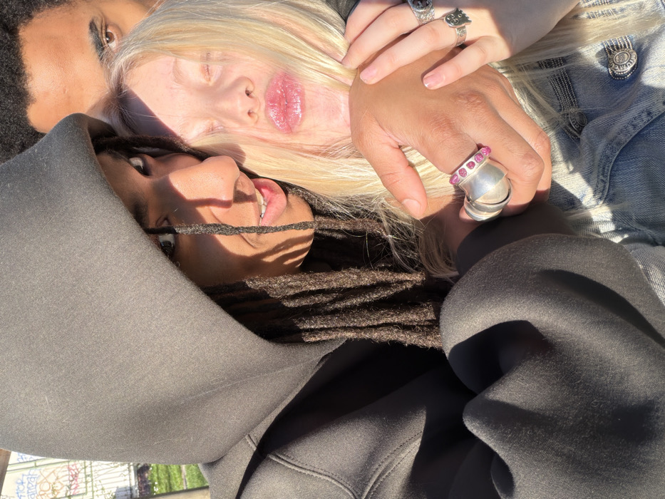 A close-up, outdoor portrait in Paris, France, features three individuals under direct, bright sunlight. In the foreground, an individual with dark dreadlocks, wearing a dark hooded sweatshirt with the hood up, gazes towards the camera with a slight smile. Their right hand, adorned with a large silver-colored ring on the ring finger and a silver-colored ring with multiple pink gemstones on the middle finger, is interlocked with the left hand of another individual. This second individual, with blonde hair and fair skin, puckers their lips towards the camera, with eyes partially closed due to the sun. They wear a light blue denim jacket and display two silver-colored rings on their left hand: one with a skull motif on the ring finger and another with an abstract design on the middle finger. A third individual, with dark, short curly hair, is partially visible in the upper background, positioned behind the other two, with only their eyes and forehead discernible, also looking towards the camera. The background consists of a light-colored fence or railing, green foliage, and faint, light-colored graffiti on a wall or panel, suggesting an urban outdoor setting. The specific location within Paris is not identifiable from the visible background elements.