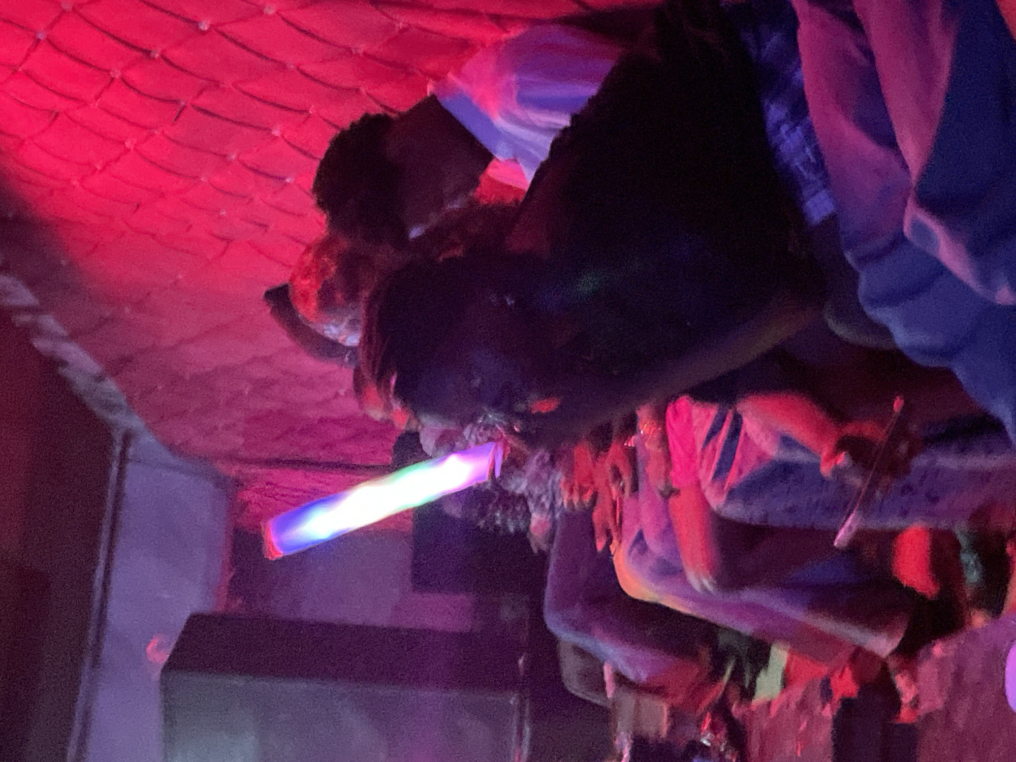In Durumi, Nigeria, an image captured in low light, likely a dimly lit establishment, shows a group of people gathered. The dominant color palette is deep reds and purples, with a bright, multi-colored light stick held by an individual in the foreground. The person holding the light stick is looking downwards and to the side, with their face partially obscured. Other individuals are seated around them, some with their backs to the camera, suggesting a casual social setting. The background features a textured, padded wall bathed in red light. The overall impression is one of a lively, possibly nighttime gathering.