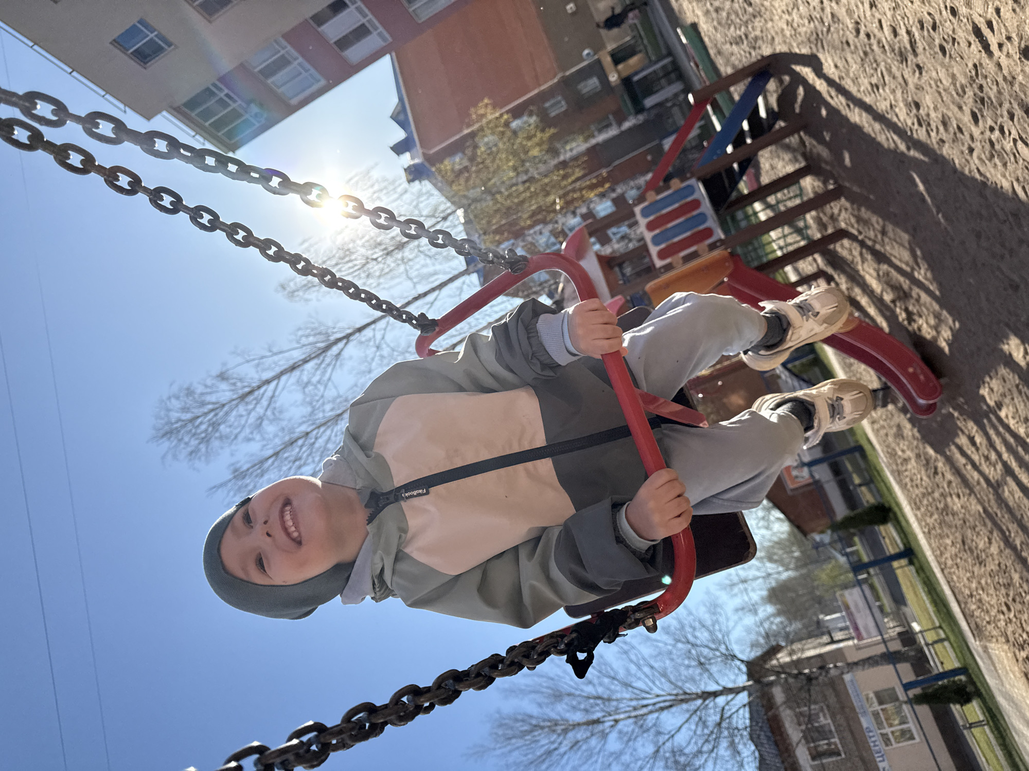 A young child is on a swing set in what appears to be a playground in Chaikovsky, Russia. The child, wearing a grey hat and a two-toned jacket, smiles at the camera while holding onto the red swing handles. The swing is suspended by chains, and the blue sky and sunlight are visible behind the chains. In the background, there are multi-story buildings and playground equipment, typical of an urban environment. The ground surface appears to be sand or a similar material.