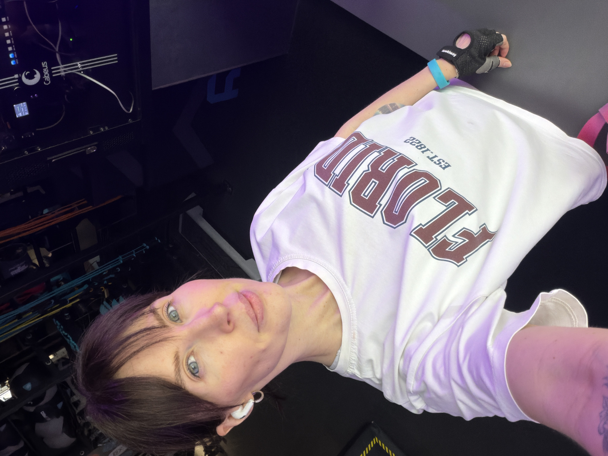 A young person with short dark hair and bright blue eyes takes a selfie in an indoor setting. They look directly at the camera with a neutral expression. They are wearing a white t-shirt featuring "FLORIDA EST.1822" in maroon and white lettering (mirrored due to the selfie). On their right hand, they wear a black fingerless gym glove with "uniktway" visible on it, along with a light blue wristband. A white earbud is visible in their left ear.
The background on the left reveals technical equipment, possibly a server rack or 3D printer enclosure, with numerous white cables, various indicator lights (green and blue), and the brand name "Cabeus" clearly visible on a black panel. The right side of the background is predominantly dark, suggesting a wall or floor, with faint blue patterns and a small section of yellow and black striped tape at the bottom. The scene is illuminated by artificial lighting, casting a subtle purple/pink hue on the person and their clothing. The location of the photo is Perm, Russia.