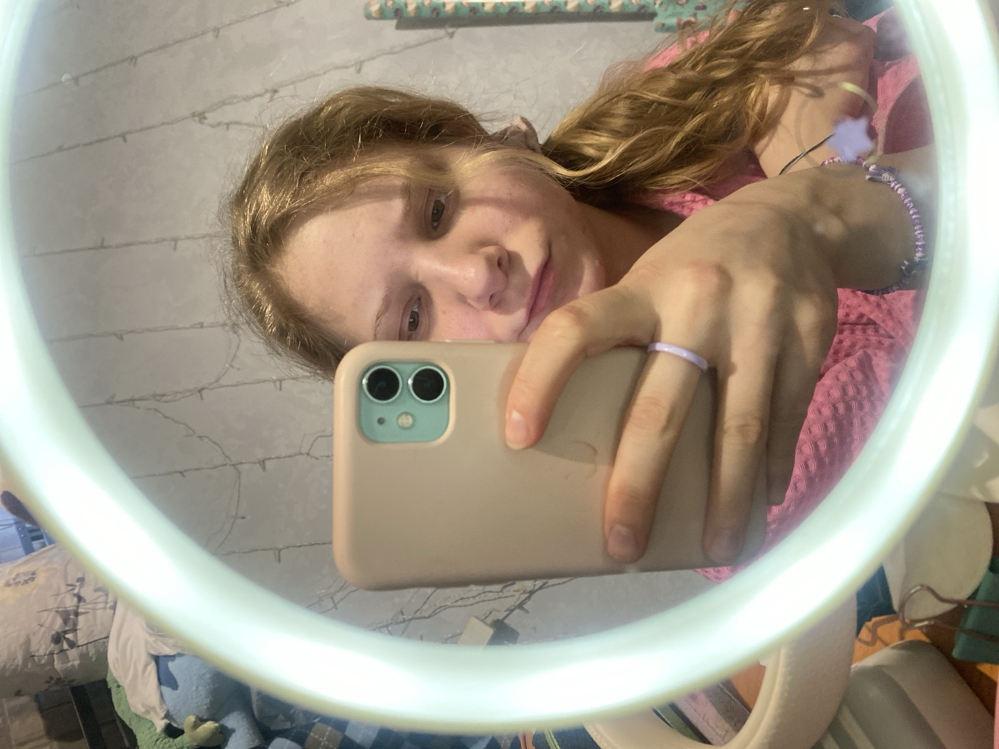 A young, fair-skinned person with light brown hair is holding a light tan iPhone in front of their face, presumably to take a selfie in a mirror. The phone has a teal green camera surround. The person is wearing a pink, textured shirt and a purple ring on their finger, as well as a beaded bracelet on their wrist. The mirror has a bright white ring light around it, and the background shows a cluttered bedroom with decorative string lights on the wall. The exact location in the city of Chaikovsky, Russia cannot be determined from the image alone.