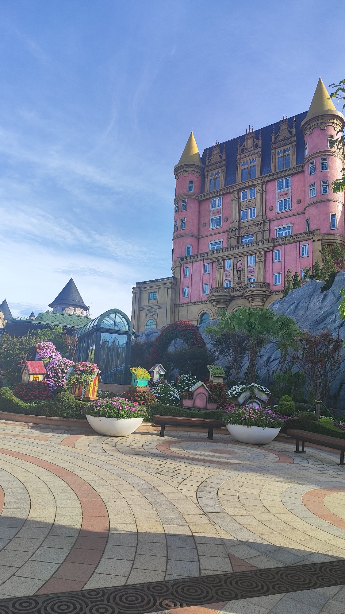 This image captures a whimsical architectural scene, likely part of a themed attraction in Đà Nẵng, Vietnam. Dominating the right side is a vibrant pink, multi-story building with pointed gold turrets, resembling a fairytale castle. The building features numerous windows and ornate architectural details. In front of this structure, a lush garden is meticulously arranged, showcasing an abundance of colorful flowers and small, whimsical houses that appear to be decorative installations rather than functional dwellings. A glass-domed structure and another building with a dark conical roof are visible to the left, adding to the fantastical landscape. The foreground is paved with patterned stone tiles, with large white planters containing flowers, and wooden benches. The sky above is a clear blue with wispy white clouds, indicating a sunny day. The overall impression is one of a playful and creatively designed outdoor space, likely intended for entertainment or leisure.