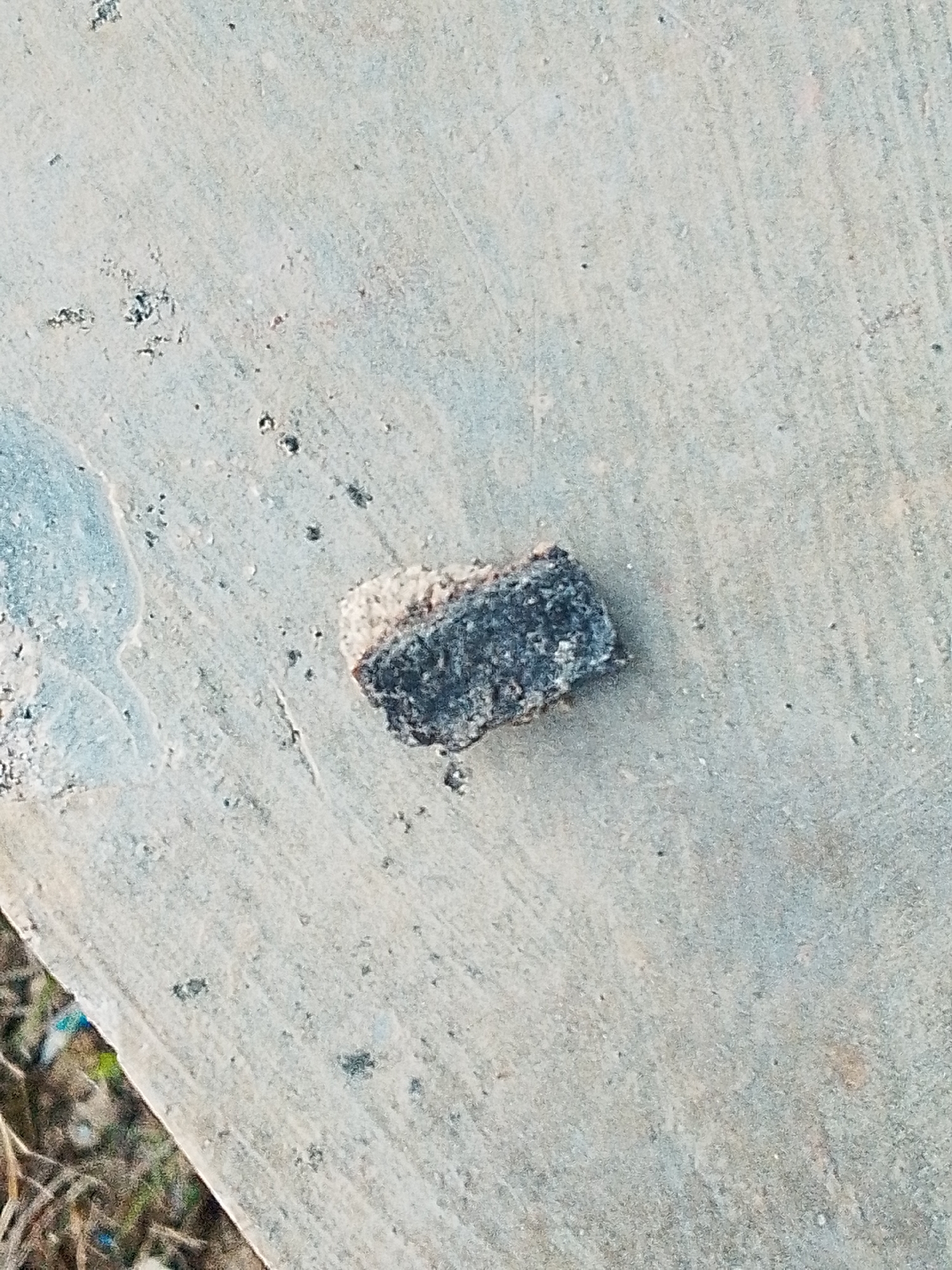 A close-up shot depicts a small, rectangular piece of dark gray material with a rough, porous surface resting on a light gray concrete surface. The material has a slightly lighter colored edge. The concrete surface exhibits subtle variations in tone and texture. Sparse debris and small dark spots are scattered on the concrete. Vegetation is partially visible in the lower left corner. The image was taken in Lhokseumawe, Indonesia. No people are visible.