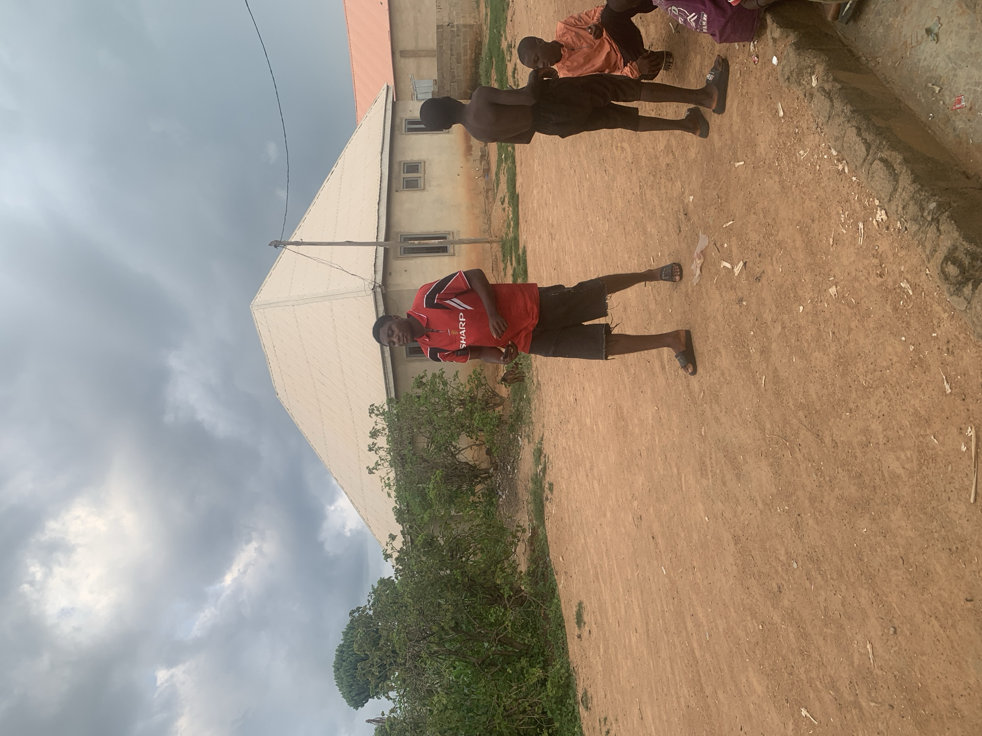 The image depicts a street scene in Sabon Tasha, Nigeria, under a cloudy sky. In the foreground, three young males are present. One individual, wearing a red and black polo shirt with "SHARP" branding and dark shorts, stands prominently in the center, looking to his left. To his right, another young male, shirtless and wearing black shorts, faces away from the camera. A third individual, dressed in an orange shirt and dark shorts, is seated or crouched to the right of the standing individuals, facing towards the left side of the frame. The ground is covered in reddish-brown dirt, with scattered debris and a concrete curb on the right side of the image. In the background, there is a building with a light-colored, A-frame roof, and further to the right, another building with an orange roof. Lush green vegetation is visible to the left of the central building. A utility pole with a wire stretches across the upper right portion of the image.