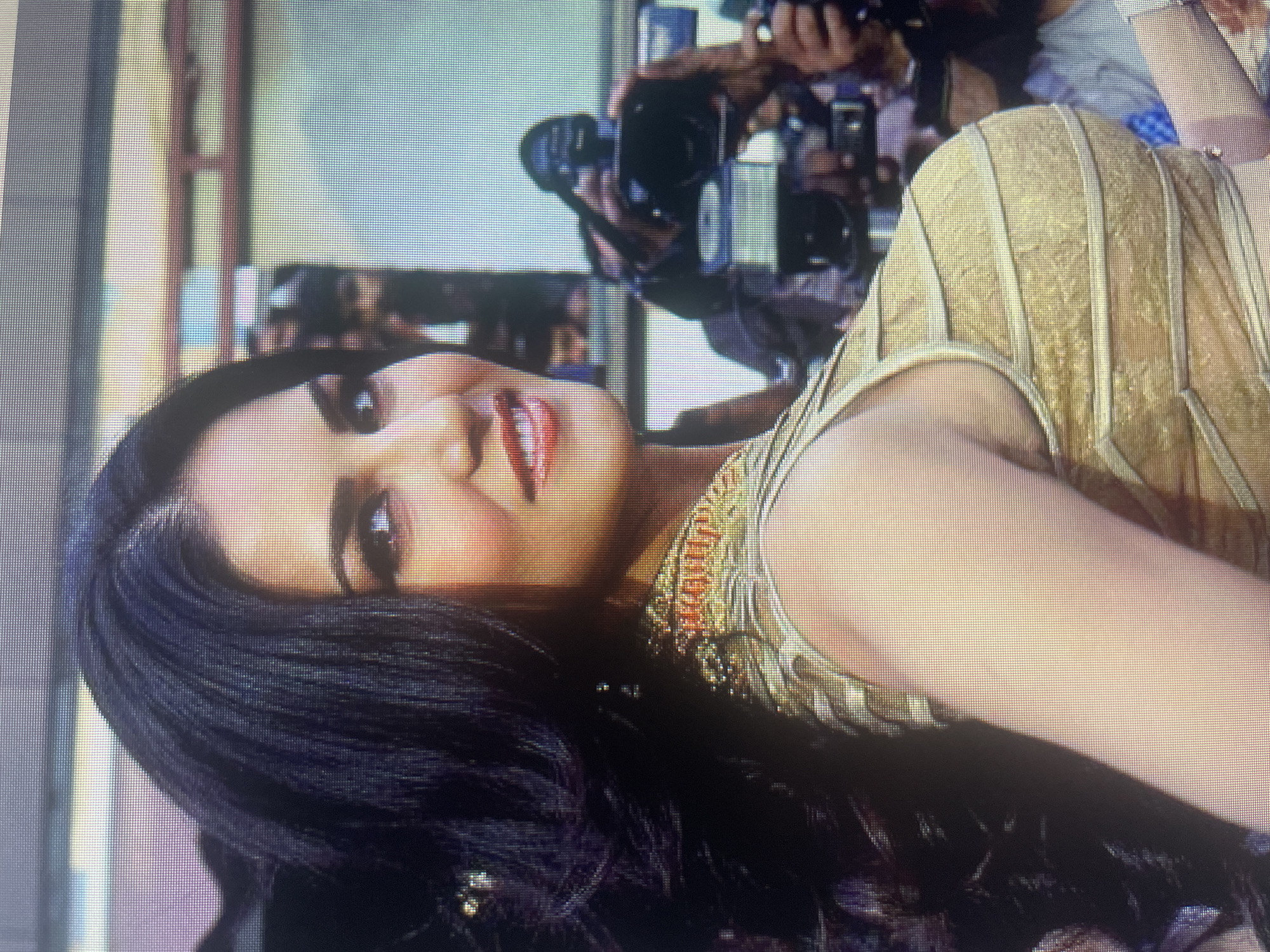 A woman with long, dark, wavy hair is shown from the chest up, angled to her right and looking towards the camera. She has full lips painted with red lipstick, and her eyes are dark. She is wearing a sleeveless, gold, intricately patterned top with ornate gold and red detailing on the straps and neckline. Her left arm is visible, resting against her side. In the background, several people are present, some holding cameras and microphones, suggesting a press event or a public appearance. The lighting is bright, and the overall scene appears to be outdoors or in a well-lit indoor venue.