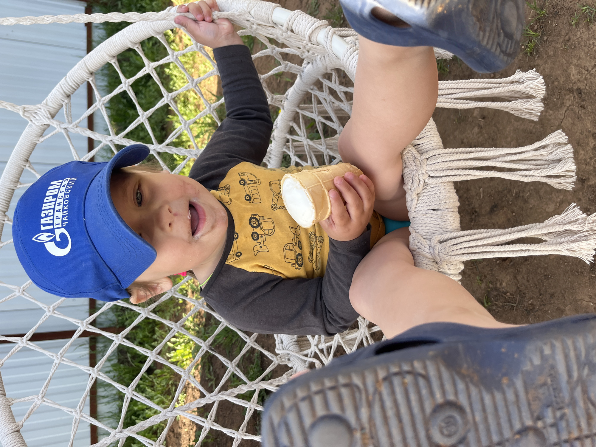 A young child, possibly a toddler, is sitting in a white macrame hanging chair. The child is wearing a blue baseball cap with the words "ГАЗПРОМ ТРАНСГАЗ ЧАЙКОВСКИЙ" printed on it, a long-sleeved shirt with yellow torso section featuring tractor images, and appears to be holding an ice cream cone. The child's face is smeared with what appears to be ice cream. A pair of blue sandals are visible in the foreground, suggesting a person is likely taking the picture from a standing position. The location is in the city of Chaikovsky, Russia.