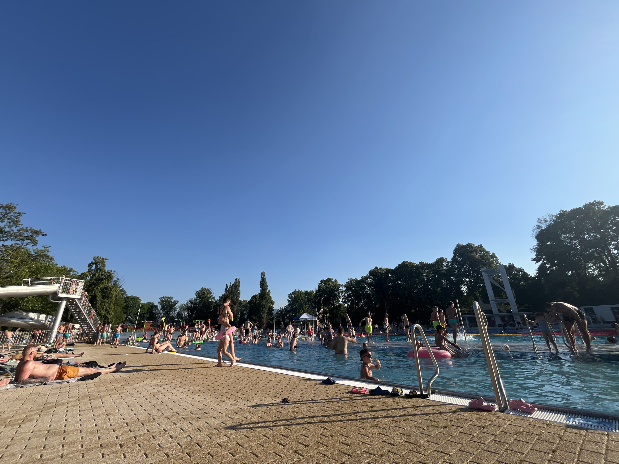 The image depicts a crowded outdoor swimming pool on a sunny day. The sky is clear and blue, suggesting fair weather. The pool area is bustling with activity, with many people swimming, wading, and relaxing by the poolside. Children are seen playing with inflatable toys, and adults are socializing and sunbathing.
In the foreground, a man is lying on a towel on the paved deck, seemingly sunbathing. Further into the scene, a large group of people are enjoying the water. Some are standing, while others are sitting or floating on inflatable rings. A diving platform with stairs is visible on the left side of the pool, with a few people standing on it. Trees line the background, indicating an outdoor recreational area, possibly a public park or a municipal pool.
The overall atmosphere appears to be cheerful and lively, with people enjoying a summer day. The sunlight casts long shadows on the paved area, suggesting it is either late afternoon or early morning. No visible text is present in the image.