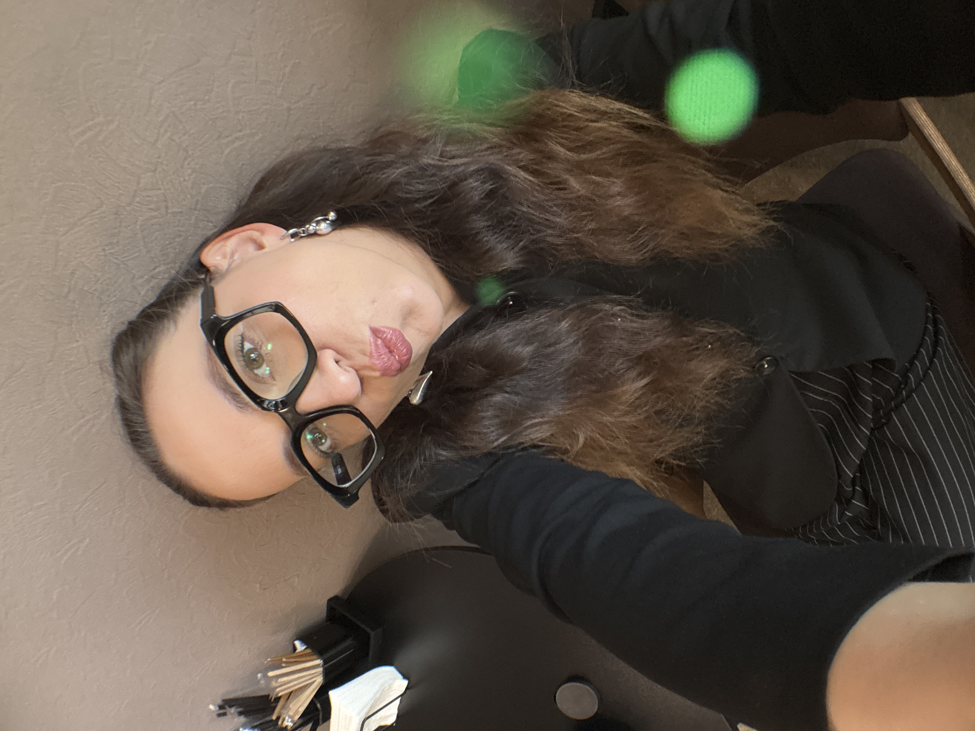 A close-up, high-angle selfie shows a woman with long, dark brown hair, wearing black-rimmed glasses and a black collared shirt. Her lips are pursed, and she is wearing a silver earring with multiple charms. In the background, a black table with a circular indentation is visible, along with a black container holding pencils. The wall behind her has a textured surface. The scene appears to be indoors, possibly in a home or office setting. There are also some green blurred lights in the foreground. The image does not provide any information or context to determine its location in Chaikovsky, Russia.