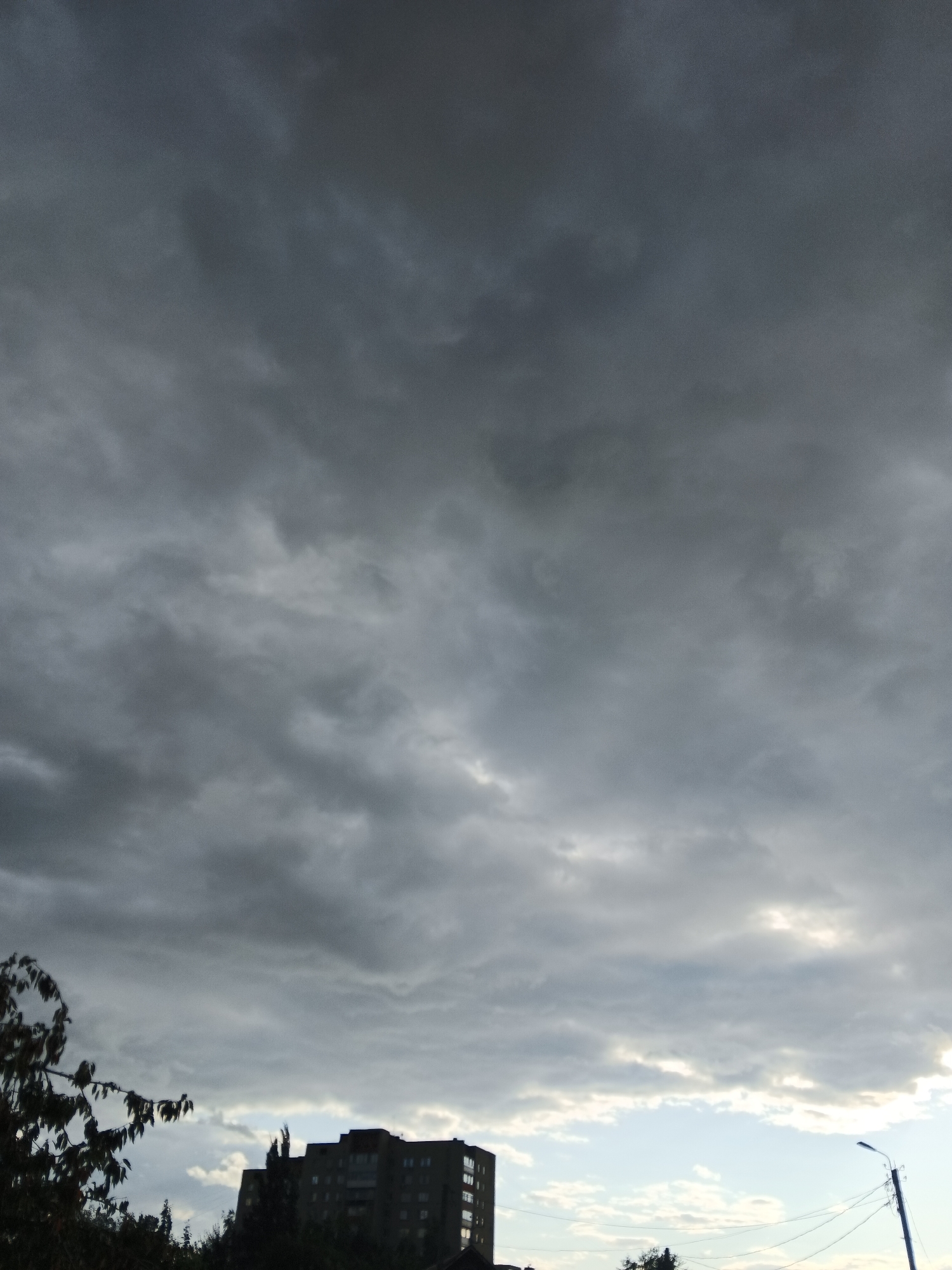 A tall, dark, overcast sky dominates the upper portion of the image, filled with thick, textured clouds. Below the dense cloud cover, a lighter sky with some breaks of blue and white clouds is visible. In the lower part of the frame, the silhouette of a multi-story building with numerous windows can be seen. To the left of the building, dark foliage from trees partially obscures the view. A streetlamp with wires is visible on the right side of the frame. The scene appears to be an urban setting under a stormy or overcast sky.