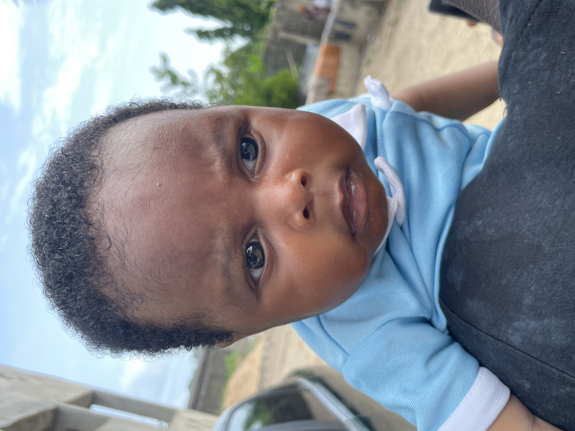 This close-up, tilted image captures a young baby looking directly at the viewer. The baby has dark skin, dark, tightly curled hair, and wide, dark eyes, with a slightly open mouth. They are wearing a light blue polo-style shirt with white trim around the collar and sleeves.
The baby is being held by an adult, whose dark-colored shoulder and upper arm are visible in the right foreground, providing support. The background is softly out of focus but reveals an outdoor setting under a bright, possibly overcast or partly cloudy sky. Green foliage from trees is visible towards the top, while concrete structures or buildings and a portion of a vehicle (possibly a car's roof and windshield) can be discerned in the lower background. The scene suggests daytime.
The baby's gaze is direct and appears curious or engaged, reflecting a moment of interaction or being photographed. The overall setting points to an outdoor location in Badagry, Nigeria. There is no visible text in the image.