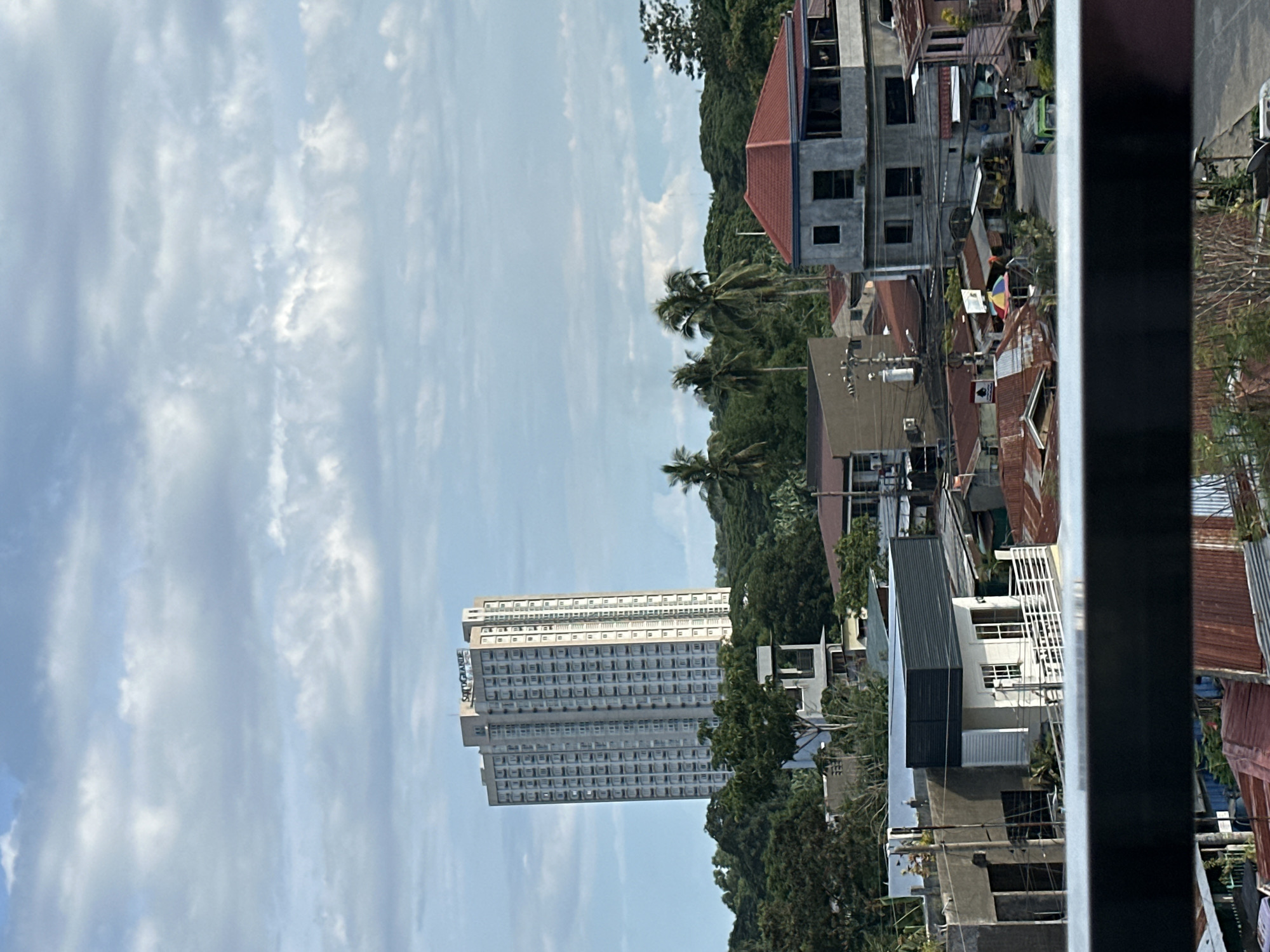 The image shows an urban landscape, potentially in Davao City, Philippines. In the foreground, a partial view of a building is framed by what seems to be a window frame or a structural beam. Beyond this foreground obstruction, the scene features a mixture of structures, including densely packed residential buildings, likely houses, with a variety of roofing materials, predominantly corrugated iron. A high-rise building with numerous windows stands out in the middle distance. The environment consists of lush greenery, including trees, which intermingle with the buildings, suggesting a setting that blends urban development and natural landscape. The sky overhead is characterized by a mixture of blue sky and scattered clouds, indicating partly sunny conditions. No people are explicitly visible in the image.