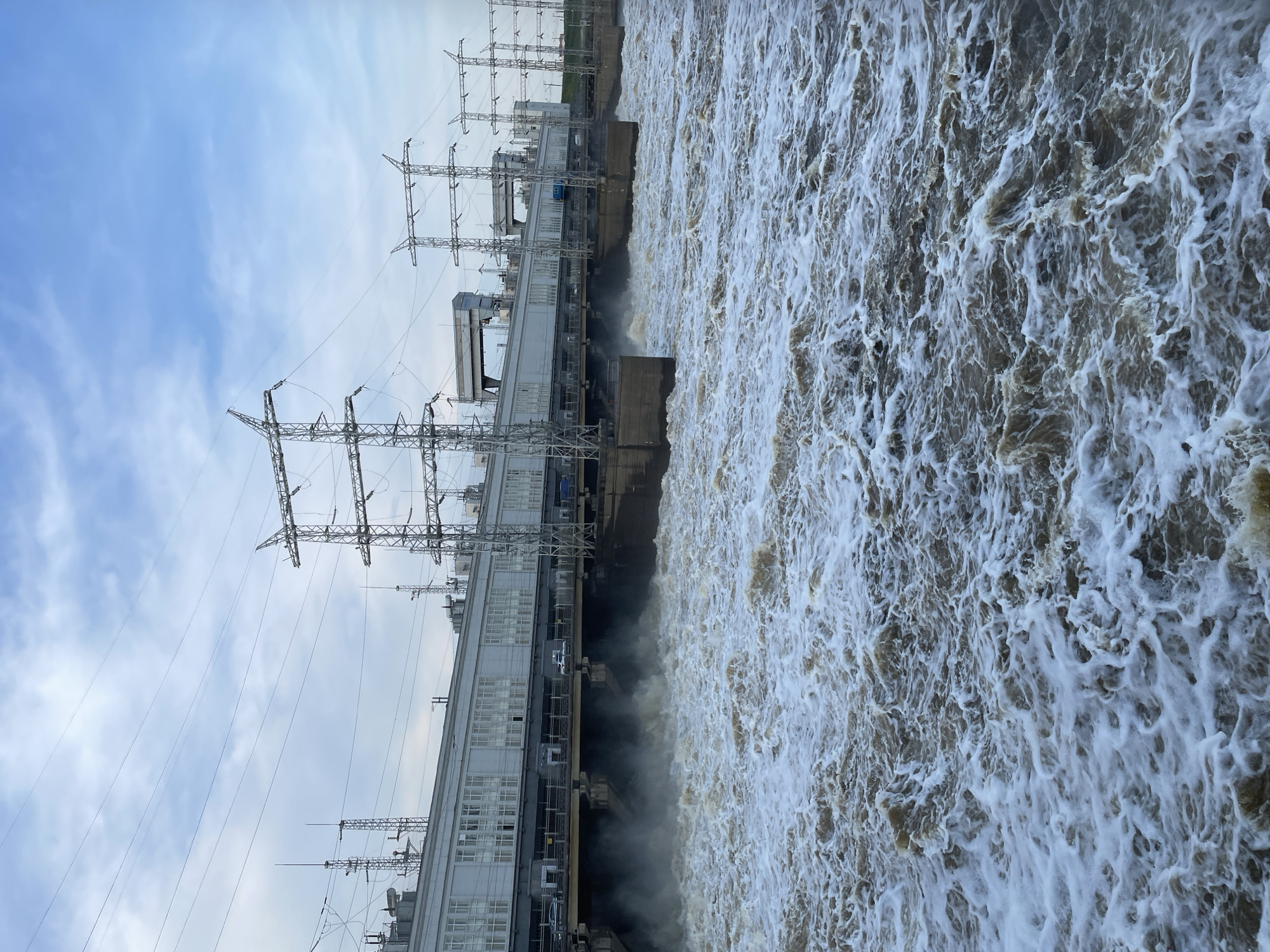 The image shows a hydroelectric power plant in Perm, Russia. The dam is releasing a large amount of water, creating a frothy, turbulent surface. The plant's architecture is visible, including the building structure and electrical transmission towers. The sky is overcast.