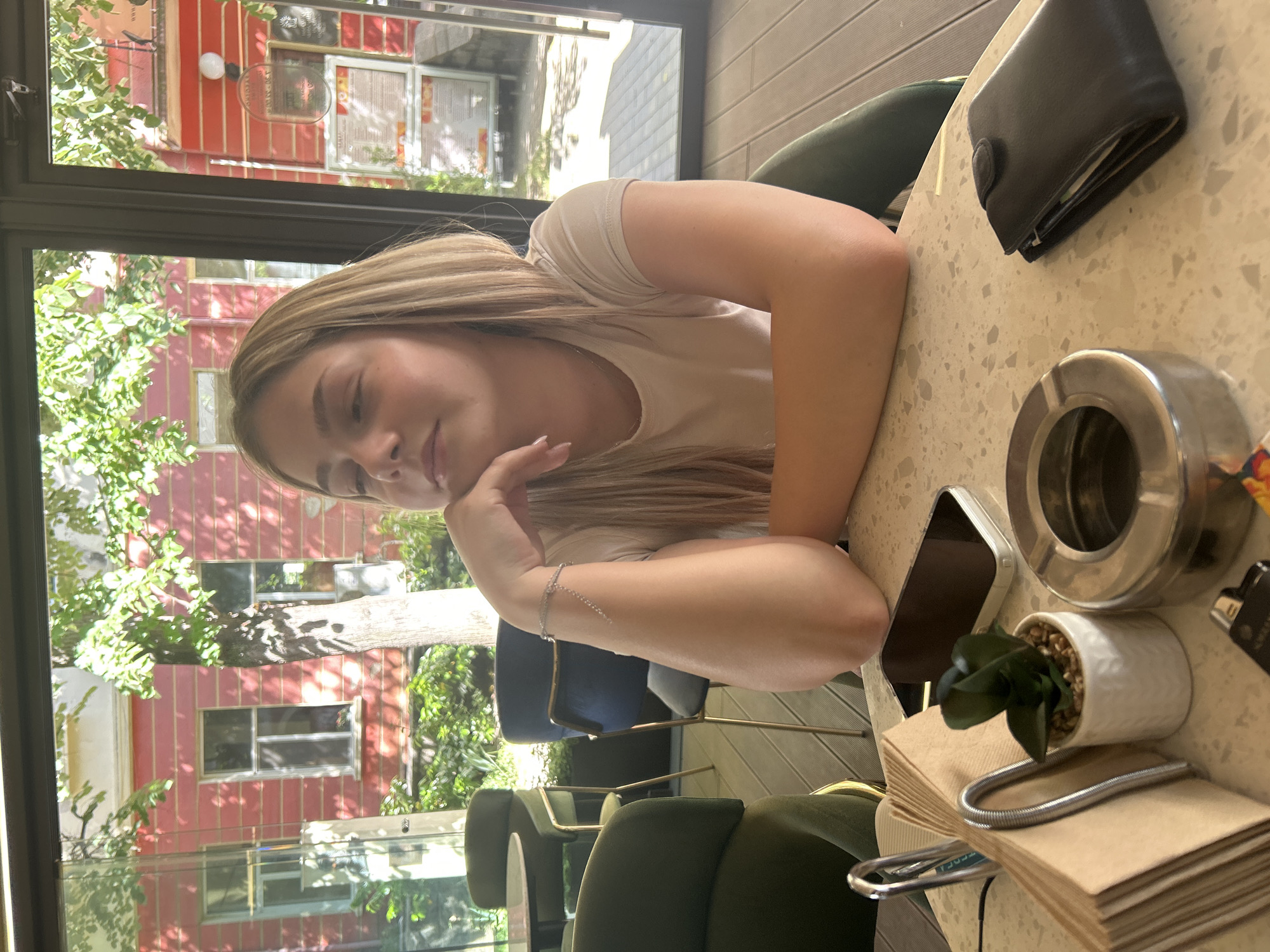 A young woman is seated at a table in an establishment, likely a cafe or restaurant. She is fair-skinned with long blonde hair, wearing a light-colored shirt. Her left arm is resting on the table, adorned with a delicate bracelet. She is looking towards the left of the frame with a slight smile. The location appears to be Kazanlak, Bulgaria, as evidenced by the architectural style and building colors visible through the window. The window offers a view of trees and a red building with several windows. On the table, next to her, is a wallet, an ashtray, a potted plant, and a stack of napkins. A smartphone is also present on the table. The chairs are upholstered in a green material.