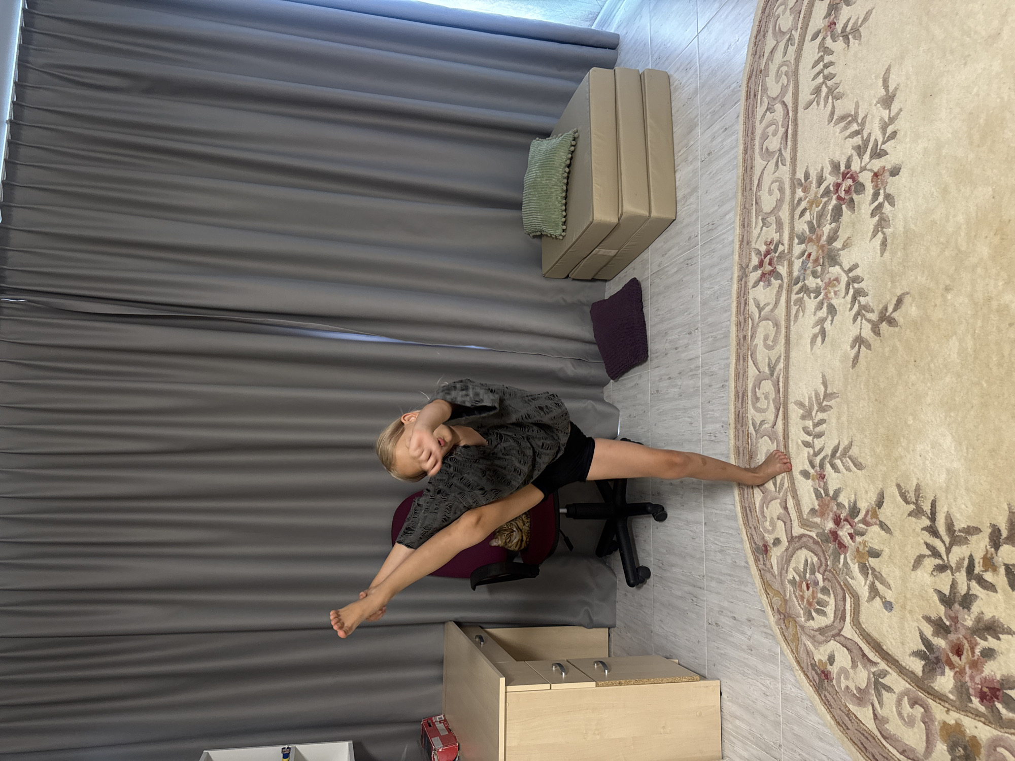 In a room with gray curtains on the left, a young person is doing a split while seated on a desk chair. They are wearing a gray patterned shirt and black shorts. Their right leg is extended forward, and their left leg is bent and tucked underneath the chair. Their right arm is stretched towards their right foot, and their left arm is bent, with their hand covering their face. A cat is visible sitting on the seat of the chair, peeking out from behind the person.
In the background, a stack of folded beige cushions is leaning against the wall. A small green cushion with pom-poms rests on top of the cushions, and a dark purple cushion is on the floor nearby. To the right of the frame, a large, round cream-colored rug with a floral pattern is visible. In the foreground, a light wood cabinet with two drawers is partially visible. The floor appears to be tiled. The image is brightly lit, suggesting it was taken during the daytime. The overall impression is of a home or studio setting where flexibility and movement are being practiced.