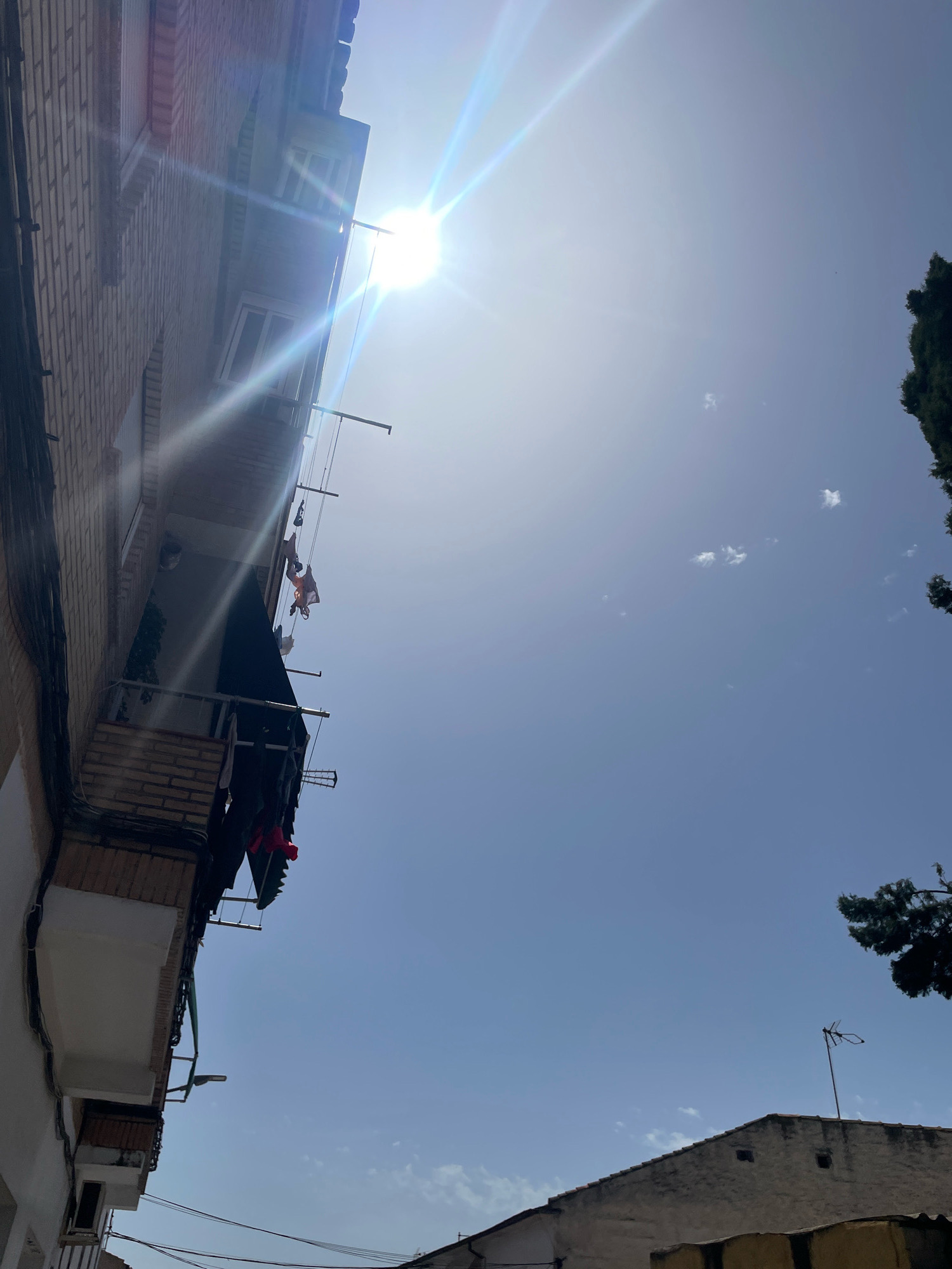 The image captures a sun-drenched view of a building facade and the sky in Atarfe, Spain. The sun is intensely bright, causing lens flare and washing out much of the sky with its glare. On the left side of the image, a brick building with white-trimmed windows and balconies is visible. Laundry is hanging from several balconies, with various garments suspended on lines. Cables run vertically along the side of the building. In the upper right quadrant of the sky, a portion of a dark green tree is visible. The lower right portion of the image shows the sloped roof of another building, with an old-fashioned television antenna atop it. The sky is a clear, pale blue with a few small, wispy clouds scattered across it. The scene suggests a warm, sunny day in an urban or semi-urban setting.