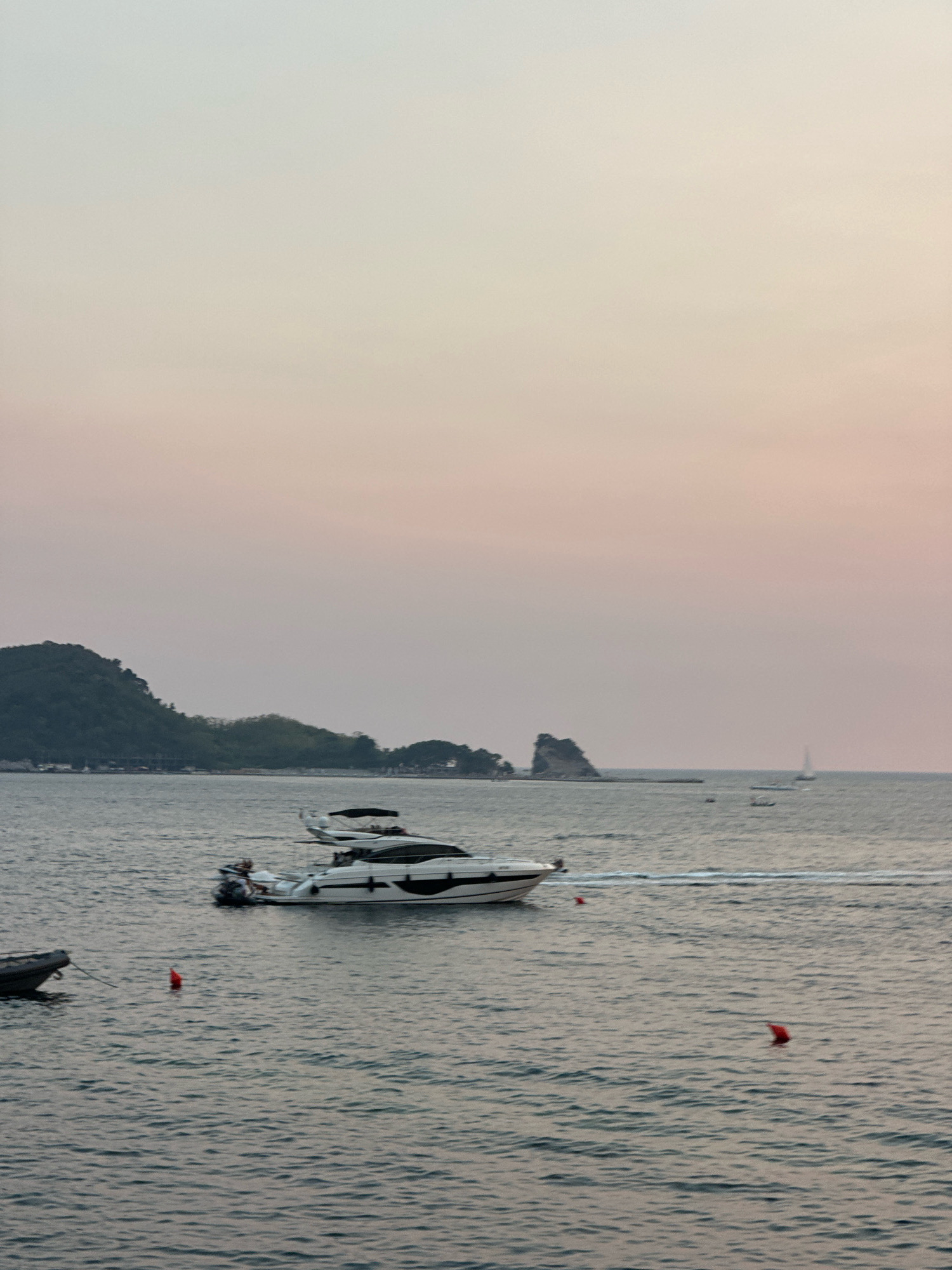 The image captures a serene coastal scene in Budva, Montenegro, likely during the late afternoon or early evening given the soft, diffused light. The sky displays a gradient of pale pinks and beiges, fading into a lighter hue towards the horizon. In the foreground, the calm, rippling surface of the Adriatic Sea dominates the lower half of the frame.
Several vessels are present on the water. A sleek, white motor yacht with black accents is the most prominent, positioned in the middle ground and appearing to be in motion, leaving a subtle wake. It has a dark canopy over the cockpit and appears to have at least two people visible on board. To the left, a smaller, dark-colored inflatable boat is tethered and appears stationary. Two red buoys are visible floating on the water, one closer to the inflatable boat and another further out.
In the background, the coastline of Budva is visible. Lush green hills and mountains form a dramatic backdrop, with some scattered buildings peeking through the foliage along the shore. Further out to sea, a solitary sailboat with a white sail is a distant spek against the horizon. Rocky outcrops jut out from the sea near the coastline, adding a rugged texture to the landscape. The overall atmosphere is one of tranquility and natural beauty.