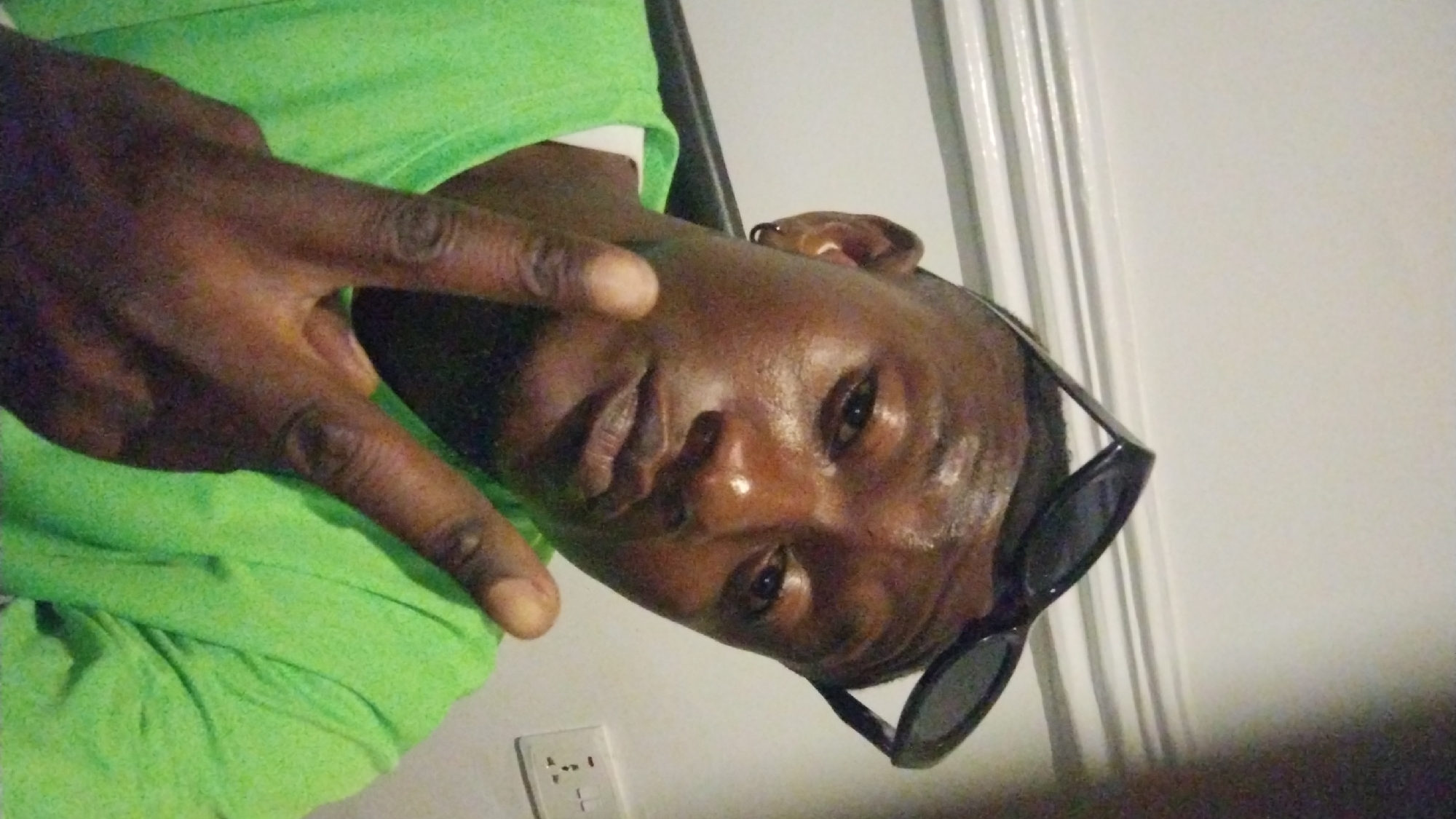An angled, close-up selfie shows an individual, likely a male with dark skin, looking directly at the camera. The image is rotated counter-clockwise. The person is wearing a bright lime green short-sleeved t-shirt. Black sunglasses are pushed up onto their head, resting above their left ear.
The person's left hand is raised prominently in the foreground, making a "peace" sign (V-sign) with the index and middle fingers extended. Their facial expression is neutral to relaxed, with highlights on the forehead and cheek suggesting indoor lighting. The individual has very short hair or a shaved head.
The background features a plain white wall with vertical architectural details, possibly paneling or a door frame, visible on the right side. A white electrical outlet with multiple sockets is mounted on the lower left portion of the wall. The scene suggests an indoor setting, consistent with a room in Jos, Nigeria. There is no visible text in the image.