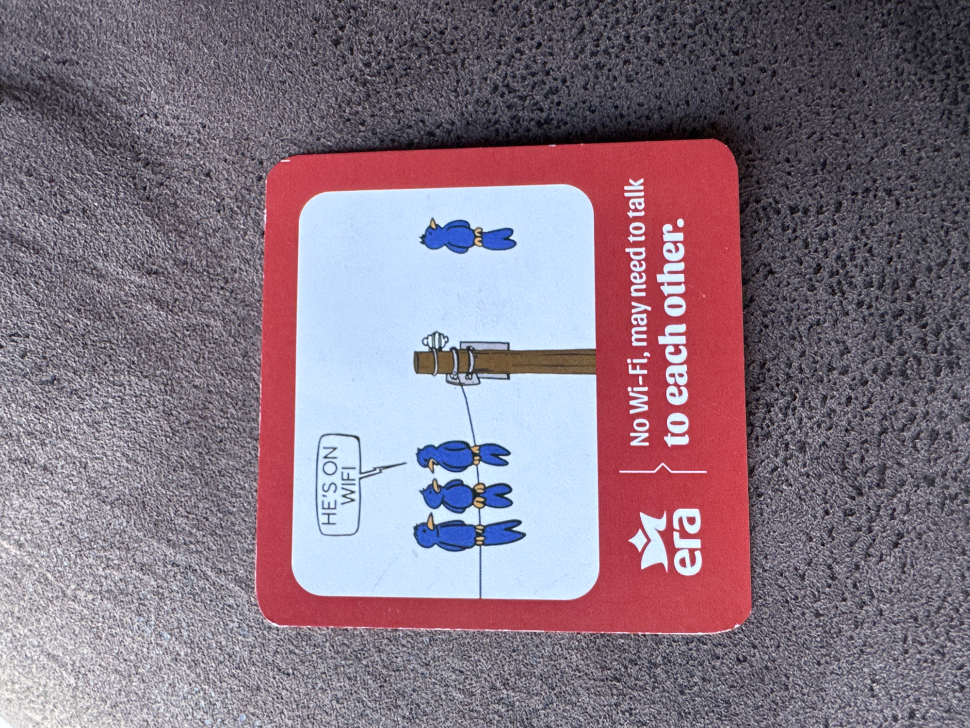 The image shows a red and white ERA Realty business card with a cartoon. The cartoon depicts a wooden telephone pole with four blue birds. Three of the birds are sitting on the wire connected to the pole and one bird is perched above the pole. A speech bubble next to the birds on the wire says "HE'S ON WIFI". The business card text reads "No Wi-Fi, may need to talk to each other." The business card is placed on a mottled grey background. The location is not indicated in the image.