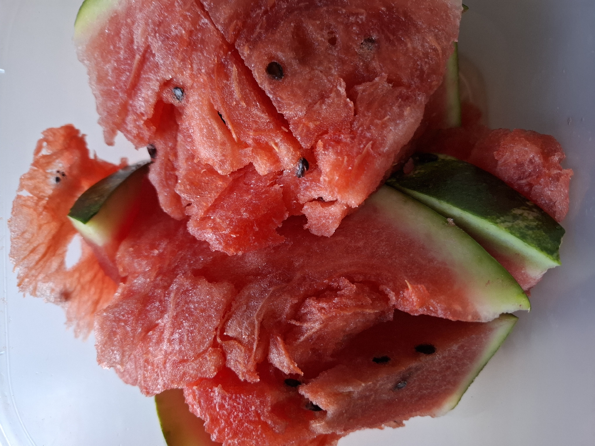 This is a close-up, overhead shot of a pile of freshly cut watermelon pieces in a clear plastic container. The watermelon flesh is a vibrant pink-red, with visible seeds. Some pieces have retained their rind, showing a green exterior with lighter green stripes. The lighting is bright, casting soft shadows and highlighting the juicy texture of the fruit. The background is a plain, light-colored surface, possibly a table. There are no people or specific landmarks visible. The scene is a simple, everyday presentation of a common fruit.
