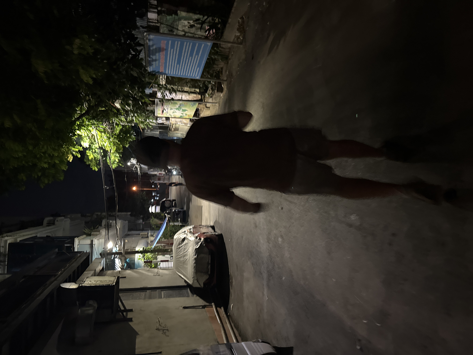 A person wearing a dark t-shirt and light-colored shorts is walking away from the camera down a narrow, paved street at night. The person has a dark baseball cap on. The street is lined with buildings on the left and a mix of buildings and trees on the right. A car covered with a light-colored tarp is parked on the left side of the street, and another vehicle is visible further down on the right. There is a blue sign with text in Vietnamese on the right side of the street. The area appears to be an urban environment in Hà Nội, Vietnam, likely a residential or back alley area given the tight spacing and building styles. The lighting is dim, with artificial lights casting shadows.