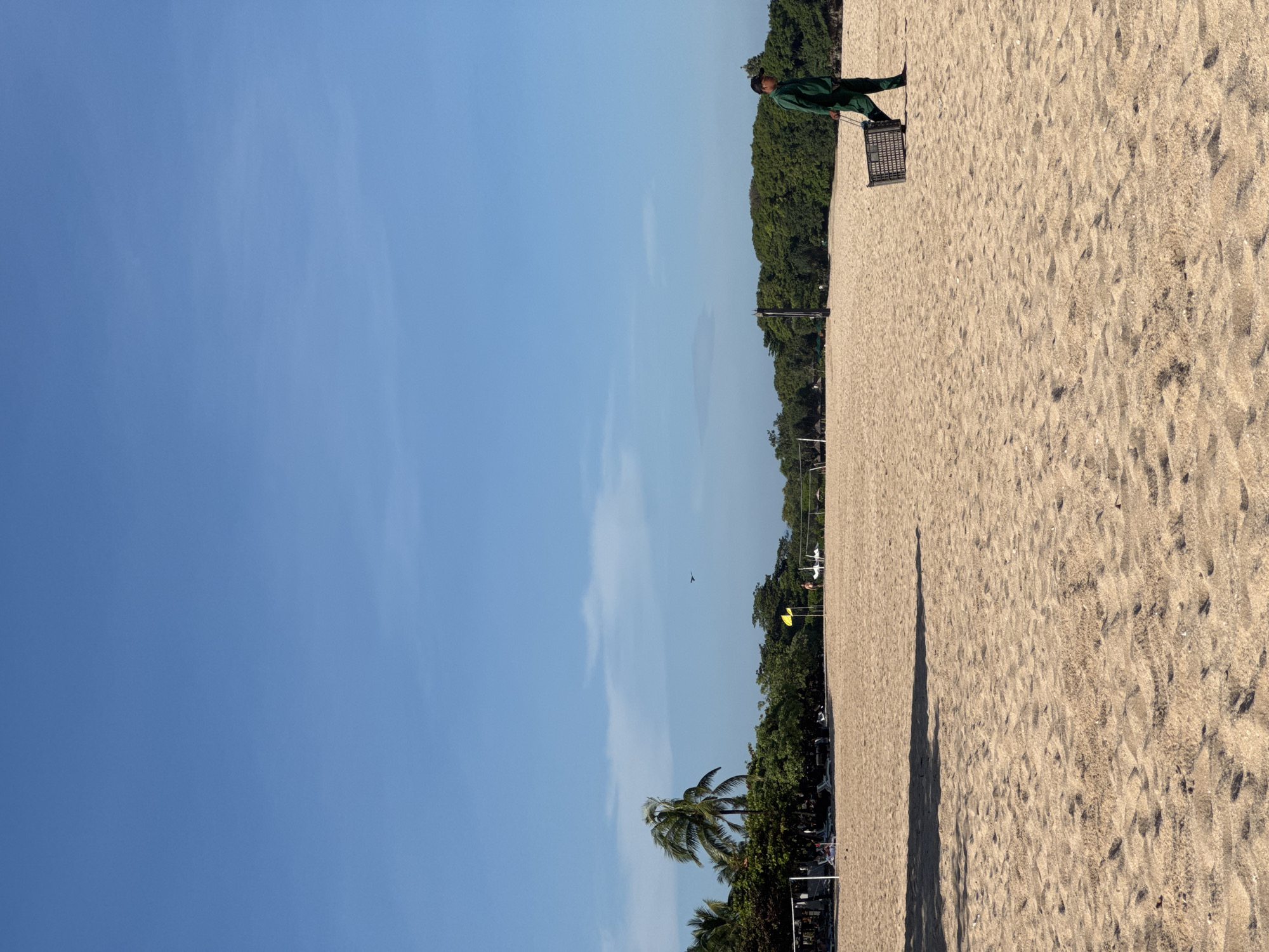 The image depicts a beach in Kuta Selatan, Indonesia on a sunny day. The sky is clear blue with wisps of clouds. The foreground features a sandy beach. A person dressed in green uniform is present at the top right side of the image, carrying a dark container, potentially for cleaning the beach. The background shows a line of dense green trees along the edge of the beach, with palm trees also visible. Beach chairs and other beach equipment can be seen near the trees. A shadow stretches across the beach.