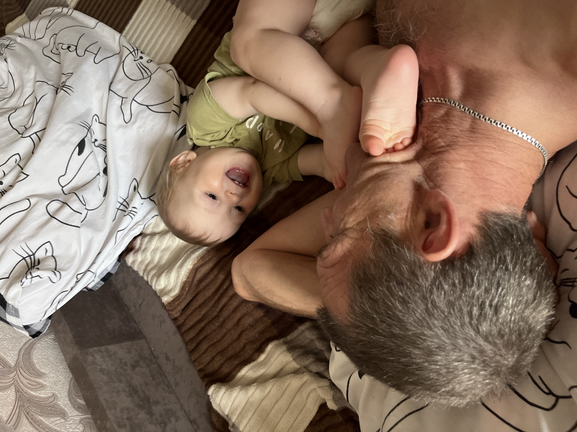 The image captures an indoor scene in the city of Noviy, Russia, featuring an adult male and a baby/toddler lying together on a bed or sofa. The adult male is positioned on his back or left side, with his head turned towards the right, mostly obscuring his face. He has short, gray hair and wears a silver chain necklace around his neck. His upper torso appears bare.
The baby, with light-colored hair, is lying on their back, positioned partially on top of the adult male's lower torso or legs. The baby wears a light green short-sleeved t-shirt with the word "HAPPY" visible across the chest. The baby's mouth is open in a wide smile, revealing teeth and tongue, indicating amusement or joy. The baby's bare feet are bent and rest directly on the adult male's neck and upper shoulder area.
The immediate surroundings include white bedding with black outline illustrations of cartoon cats or rabbits, partially covering the baby and extending to the top of the frame. Beneath both individuals is a textured brown blanket or throw with alternating light and dark brown striped sections. To the left, a dark brown, possibly upholstered, surface is visible, adjacent to a light-colored wall featuring a subtle, textured pattern. The lighting in the scene suggests an interior environment without harsh direct light.
