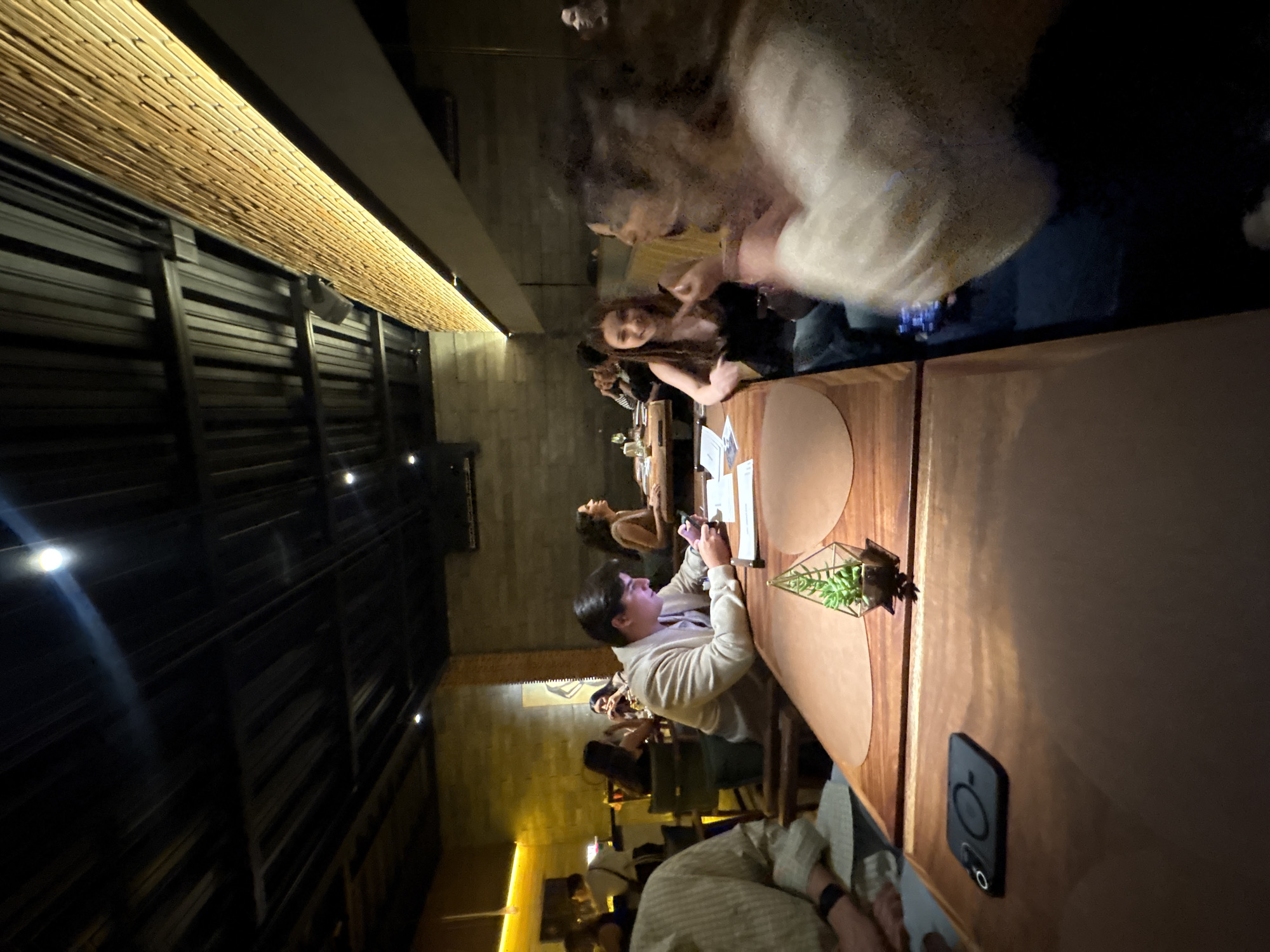 A brightly lit, indoor scene in San Andrés Cholula, Mexico shows several individuals seated at a wooden table. A man wearing a light-colored jacket is seated at the table, looking toward the center. There are other people seated along the table. The table contains plates, utensils, and a small geometric terrarium. A phone sits face up next to the table. The architecture includes a thatched or woven ceiling detail and vertical black slats lining the upper portion of the walls. The lighting is warm, creating a casual and intimate atmosphere.