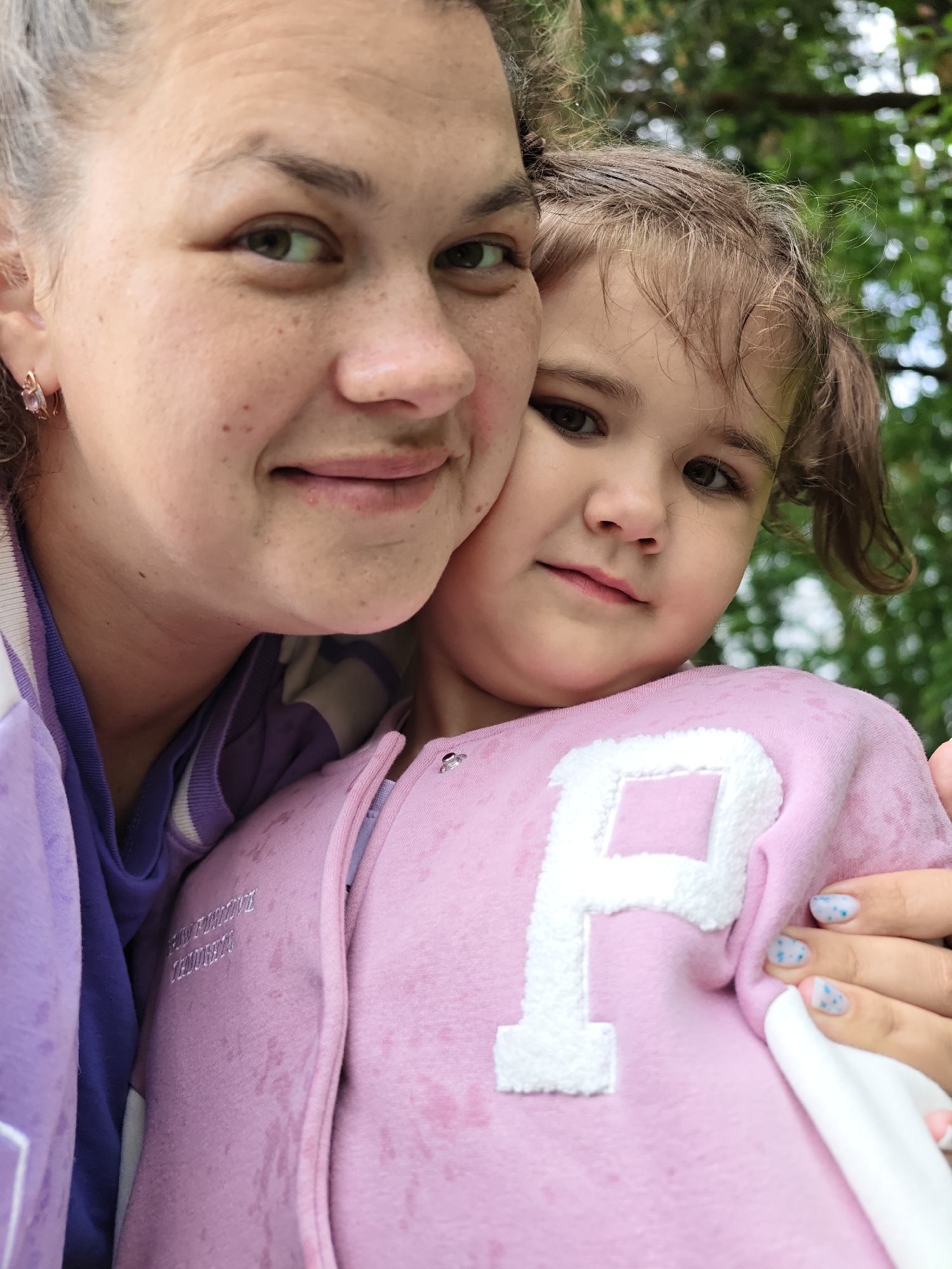 A close-up outdoor portrait features two individuals, an adult and a young child, taken in Chaikovsky, Russia. The adult, positioned on the left and partially cropped, has light skin with visible freckles, green-brown eyes, and light brown hair pulled back. They wear a purple and white striped top under a solid purple jacket and have a small, light-colored earring in their visible ear. A subtle smile plays on their lips as they look towards the camera.
The young child is held close by the adult, prominent on the right side of the frame. They have dark brown eyes, light brown, curly hair, and a gentle, slightly contemplative expression as they gaze directly at the viewer. The child is wearing a pink jacket or hoodie, which features a large, white felt letter "P" on the front. On the side of the pink jacket, the embroidered text "GROW POSITIVE THOUGHTS" is visible. The child's fingernails are painted light blue and white with small blue dots.
The background is softly blurred green foliage, indicating an outdoor setting, possibly a park or garden, under bright, diffused daylight. The image captures a warm, intimate moment between the two subjects.