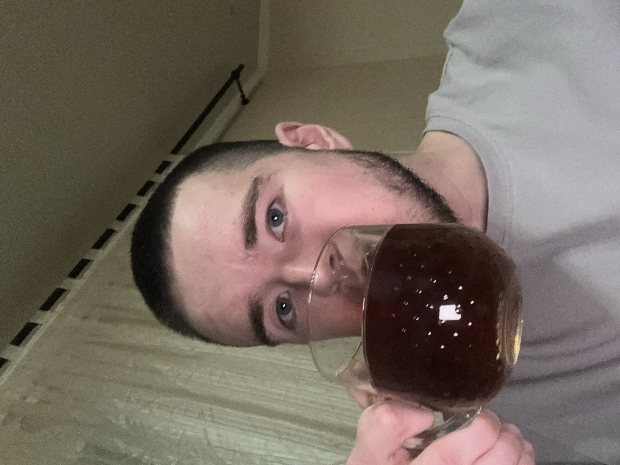 A man is holding a glass of dark liquid, possibly coffee or tea. He has short dark hair and light-colored eyes. He is wearing a light grey t-shirt. The background is out of focus but appears to be a room with light-colored walls and flooring. It is not possible to determine if this image was taken in Kazan, Russia without additional information.