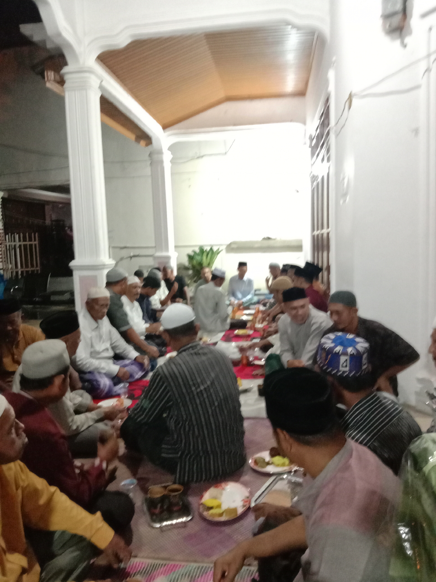 The image shows a gathering of approximately 20-25 Muslim men sitting on mats on the floor, seemingly in an indoor/outdoor space. The location is likely Lhokseumawe, Indonesia.
The men are dressed in a mix of casual clothing, including shirts, sarongs, and some are wearing traditional Muslim caps (peci). Some are seated on the floor with crossed legs, while others are in a kneeling position. Plates of food and drinks are placed on mats in front of them, indicating a meal or gathering involving food.
The setting appears to be a covered area or patio adjacent to a house. There are white pillars that support the structure, and a wooden ceiling is visible. The lighting is artificial and somewhat dim. The overall atmosphere suggests a communal gathering, possibly a religious or cultural event.