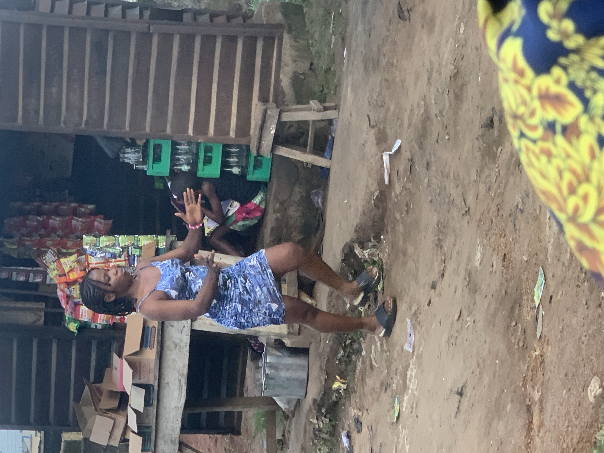 A young Black woman with braided hair walks on a dirt path in front of a wooden stall in Shagamu, Nigeria. She wears a blue and white patterned dress and dark sandals. The stall appears to be a small shop selling drinks and snacks, with bottles and packaged goods displayed. A large metal bucket sits to her left, and several cardboard boxes are stacked on a wooden table to the left of the stall. The ground is uneven and littered with debris. In the background, other people are partially visible near the stall. The overall scene depicts a typical street-side market environment.