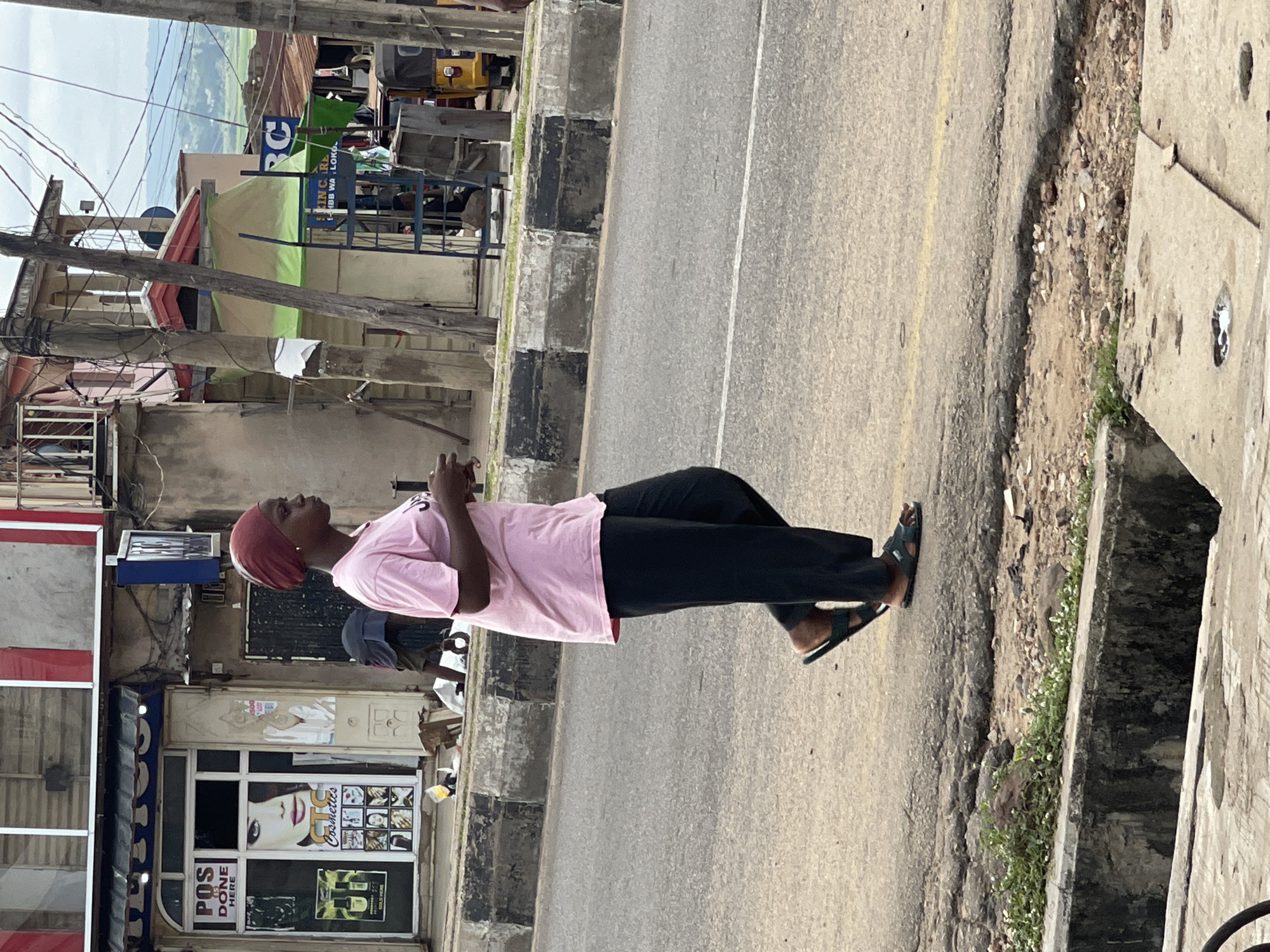A young woman wearing a pink t-shirt and black pants walks on a paved road. She has a red headscarf and is wearing black sandals. She is walking in front of a row of shops. The shops have signs that are written in English. The road is made of asphalt and has some potholes. There is a concrete curb on the side of the road, and some vegetation growing along the curb. In the background, there are buildings and trees. The sky is cloudy.