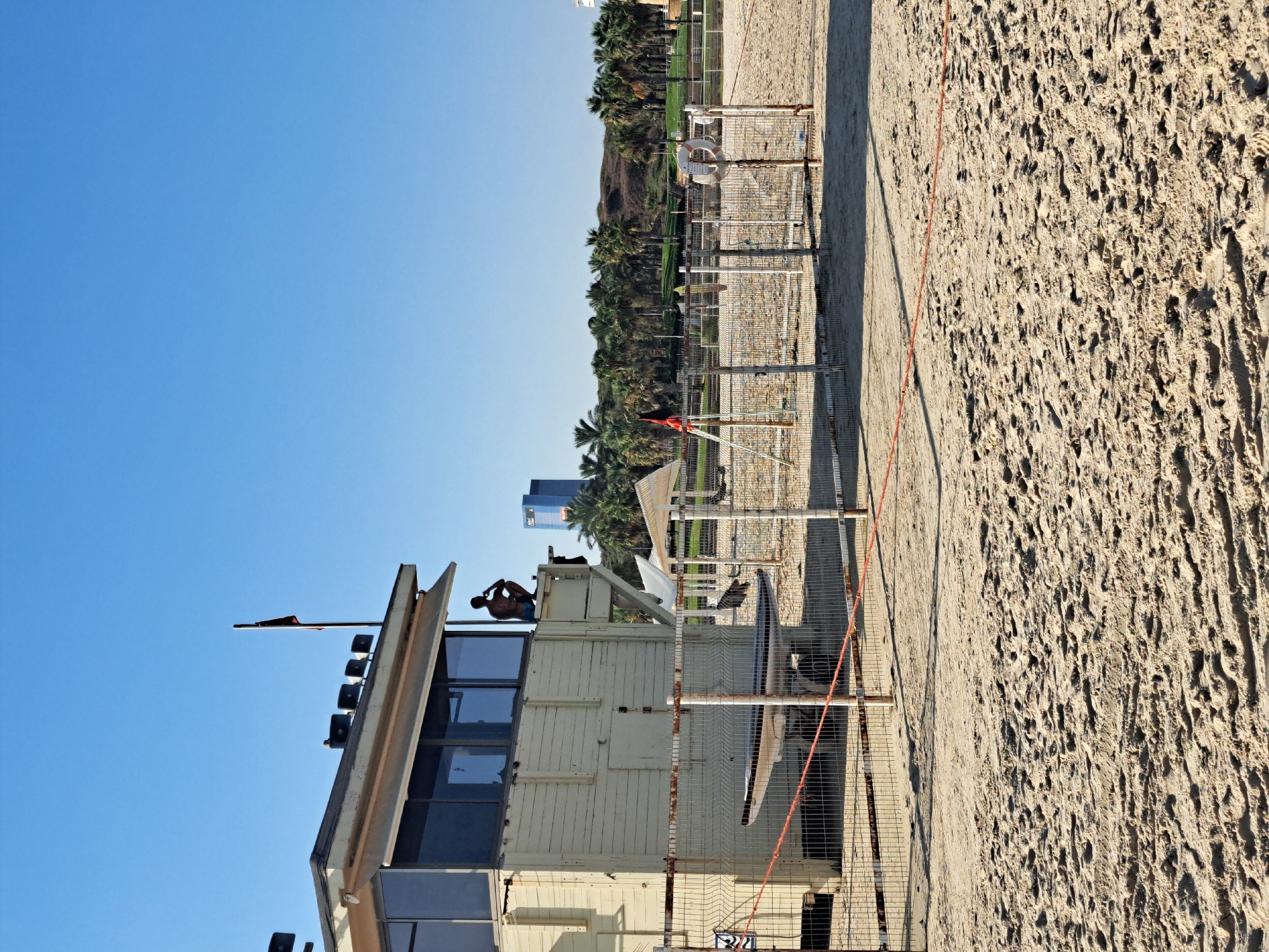 An outdoor daytime scene on a sandy beach in Herzliya, Israel, depicts a lifeguard station, a fenced-off area, and palm trees in the background. A man, appearing to be a lifeguard, is seated on the top of the light-colored, wooden lifeguard station, looking to his left. The station has glass windows and a retractable awning. A flag is mounted on a pole on top of the station. A surf-board is visible on a cart behind a metal fence. The fenced area contains a life preserver attached to a pole. In the background, a row of palm trees lines the horizon, with a hill behind them. Two modern, tall buildings are visible in the distance, with one of them being a bright blue skyscraper. The sandy ground is imprinted with footprints.