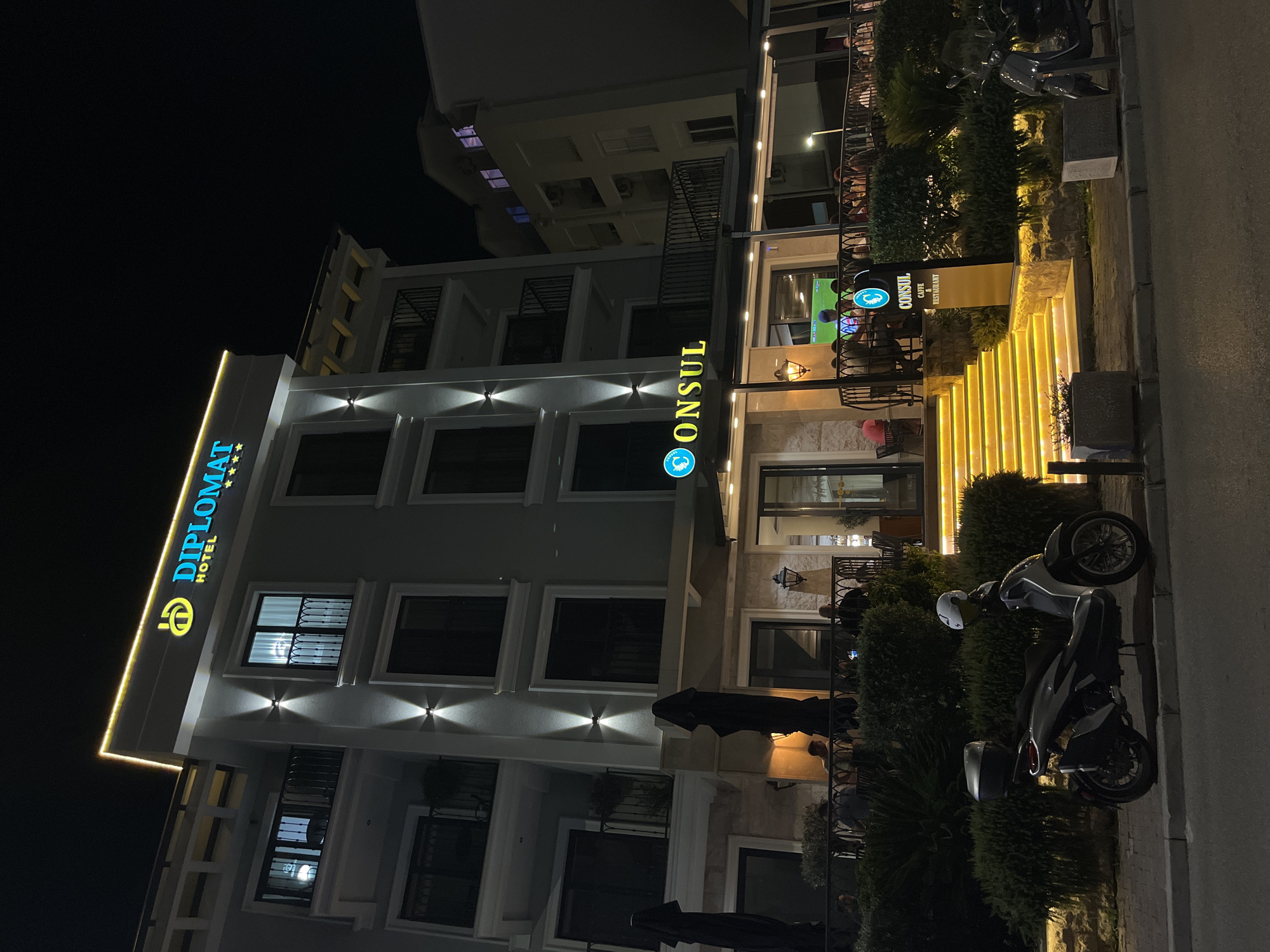This is a nighttime photograph of the Diplomat Hotel and Consul Caffe & Restaurant located in Budva, Montenegro. The hotel is a multi-story building with illuminated signage displaying "DIPLOMAT HOTEL" in blue lettering against a yellow outline. Below the hotel, the "CONSUL CAFFE & RESTAURANT" sign is also illuminated. The entrance to the restaurant features a set of brightly lit yellow stairs, flanked by hedges and outdoor seating areas. Several people are visible dining and socializing on the restaurant's terrace. In the foreground, a silver scooter is parked on the sidewalk. The scene is illuminated by artificial lights, highlighting the building's architecture and the outdoor dining space.