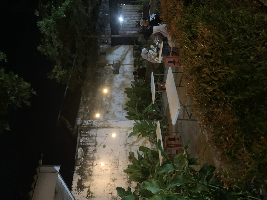 The image shows a night scene at a cafe, located in Probolinggo, Indonesia. The cafe is situated against a whitewashed building with a string of lights. There are three tables and four chairs set up on the patio. Two women are sitting at a table in the foreground, facing away from the camera. A man in a black shirt and a white baseball cap is sitting beside them. The scene is illuminated by the string lights, creating a warm and inviting ambiance. There is lush foliage surrounding the patio, adding to the tranquility of the scene.