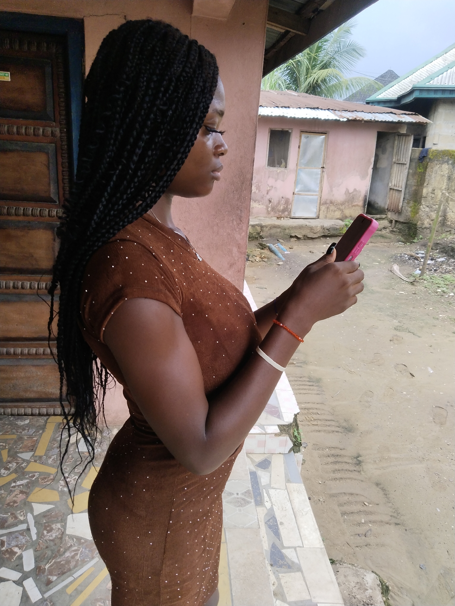 A young woman stands on a tiled porch in Uyo, Nigeria, facing right, holding a pink-cased mobile phone in both hands. She is wearing a short-sleeved, brown, bodycon dress adorned with small, reflective studs. She has long, dark braids. A white bracelet and an orange beaded bracelet are on her right wrist. Behind her is a dark brown wooden door and a pink wall. Beyond the porch, there is a sandy yard with two small, dilapidated buildings. One building is pink and the other is a greyish-blue color. The sky is overcast.
