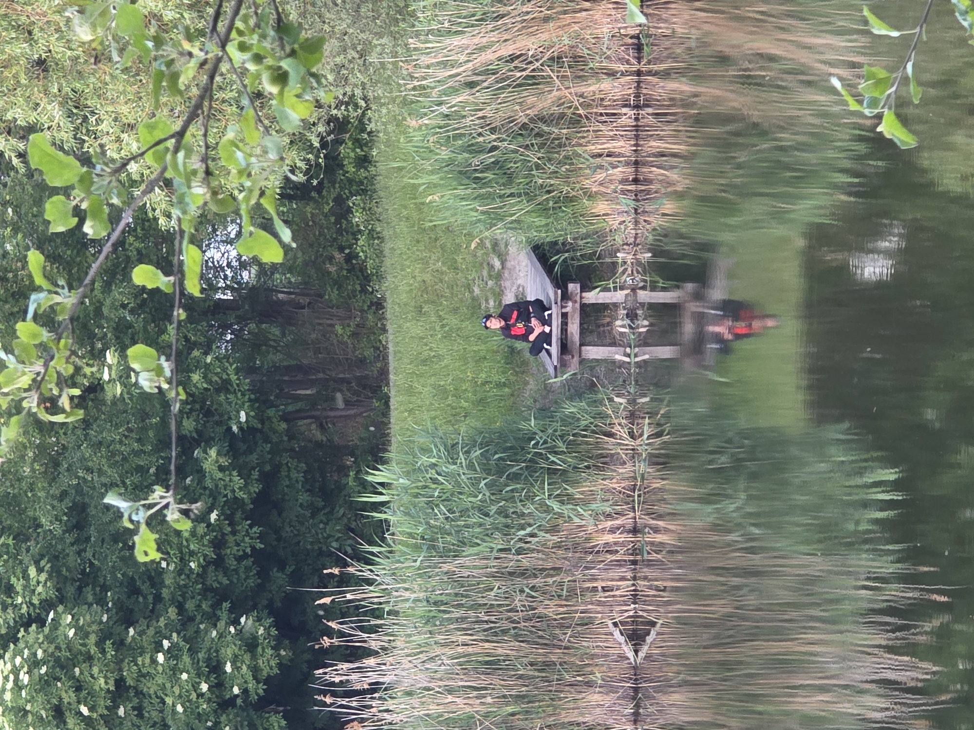 A person sits on a wooden platform extending into a body of water, surrounded by tall reeds. The person is wearing dark clothing with red accents and a dark hat. Their legs are crossed, and they are facing towards the left of the frame. The water reflects the person, the reeds, and the greenery of the background, which consists of trees and grass. The scene appears to be a natural setting, possibly a park or a nature reserve, within the city of Warsaw, Poland. The lighting suggests it is either early morning or late afternoon.