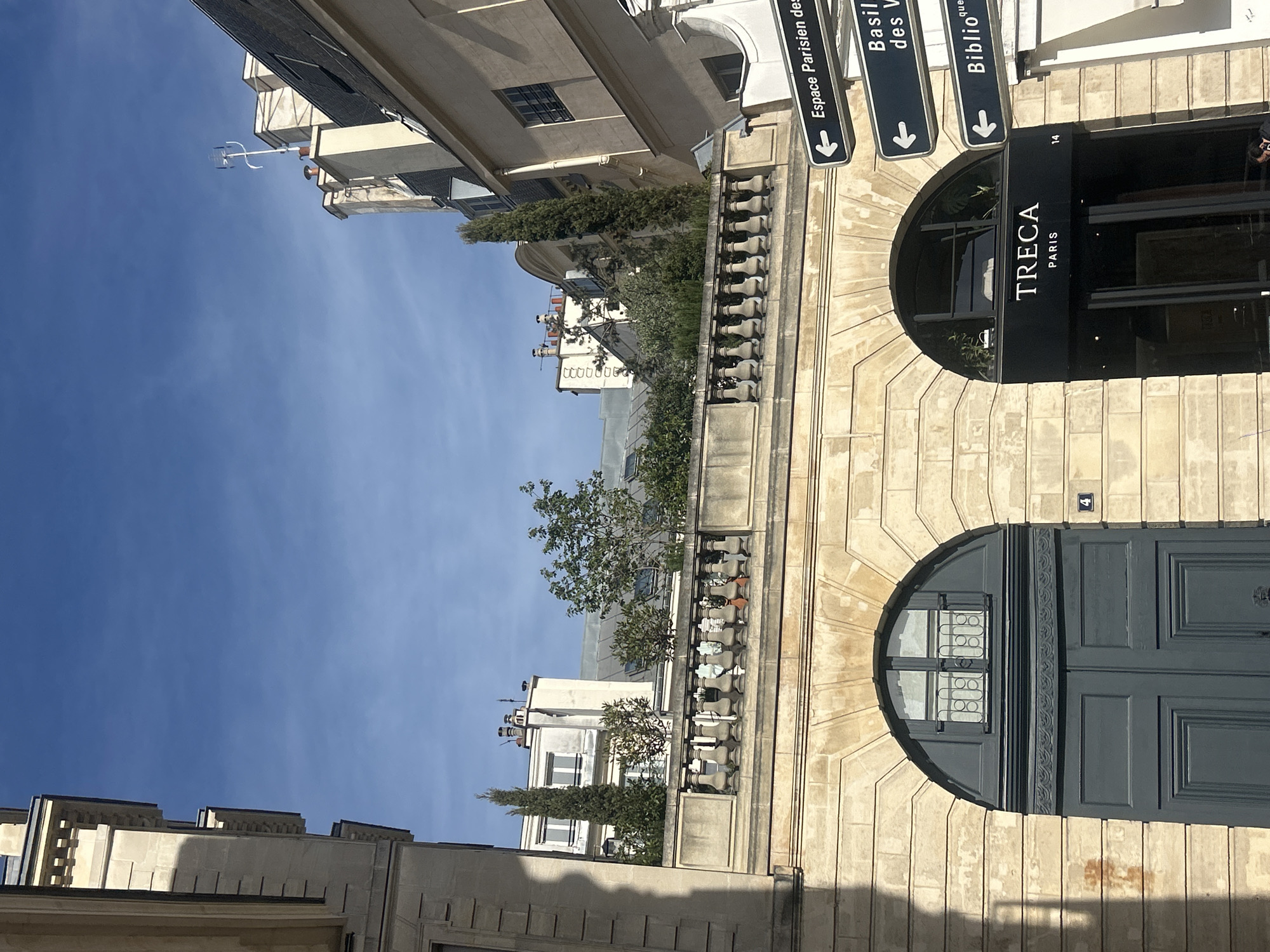 The image shows a street in Paris, France. On the right side of the image, there is a building with two arched entrances. The top entrance is the shop TRECA Paris, and there are street signs pointing towards Espace Parisien and Basilique des Victoires. The other entrance features a large gray door with a window with a decorative grill. The building is made of light-colored stone and has a balustrade with greenery behind it. In the background, there are other buildings and a blue sky.