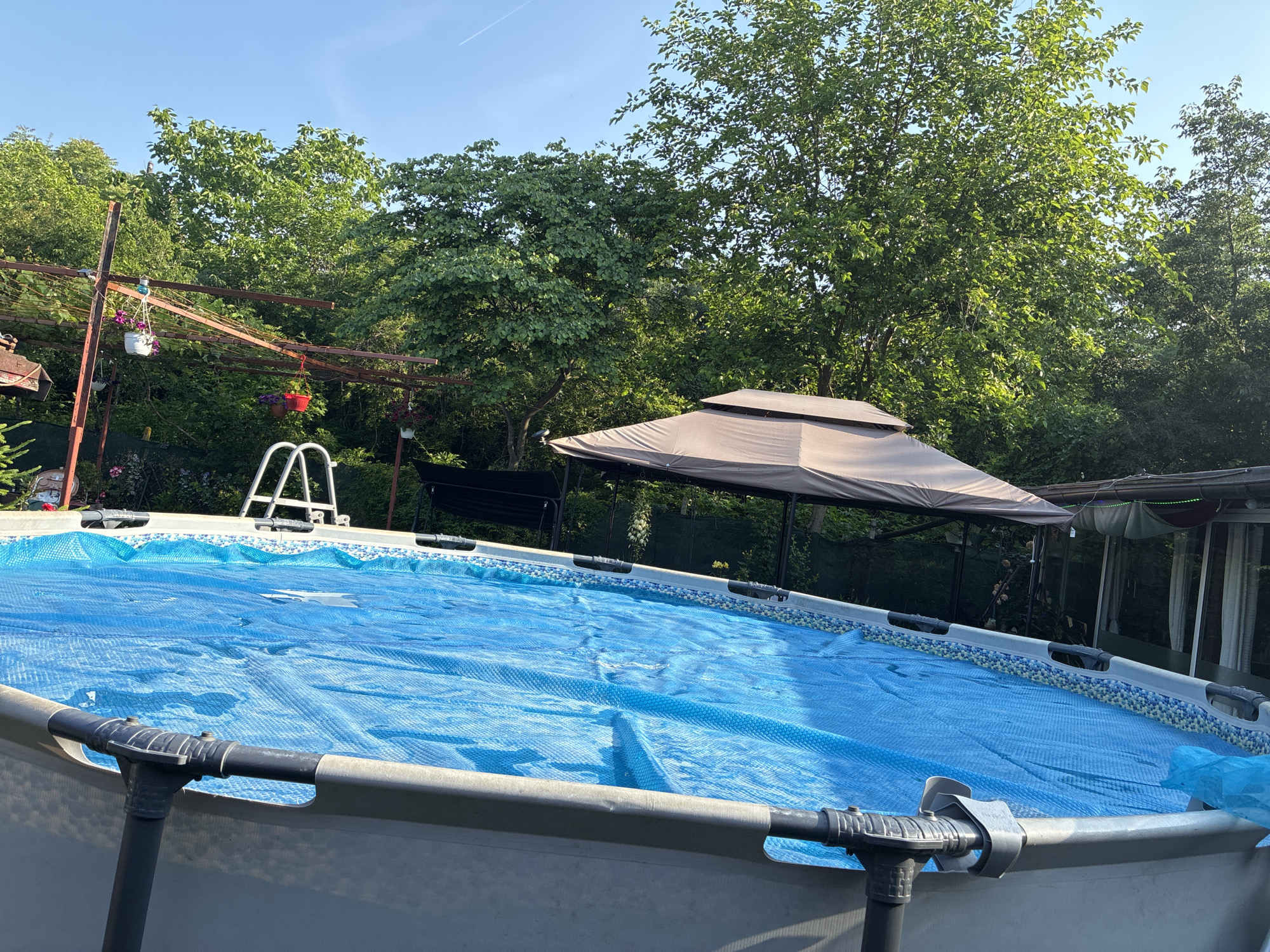 This forensic description pertains to an outdoor scene in Varna, Bulgaria, captured during daylight hours under a clear, bright blue sky with a visible aircraft contrail.
The primary subject is an above-ground swimming pool, partially visible in the foreground and mid-ground. The pool is oval or rectangular with rounded corners, featuring a light grey exterior wall and a blue-and-white patterned interior rim. A blue bubble-wrap style solar cover floats on the water's surface, covering the majority of the pool. A white metal ladder is positioned at the far side of the pool, providing access to the water.
Surrounding the pool is dense, lush green foliage consisting of numerous trees and bushes, indicating a garden or backyard environment. In the mid-ground, a brown, peaked-roof gazebo or canopy stands to the right of the pool. To the left, an aged, rusted metal pergola or trellis structure supports vines and features multiple hanging potted plants with purple flowers in white and red containers. A dark-colored outdoor swing or bench is partially visible beneath the trees behind the pool. No individuals are present in the depicted scene. The sunlight suggests a sunny day, likely during late morning or early afternoon, based on the shadows and illumination.