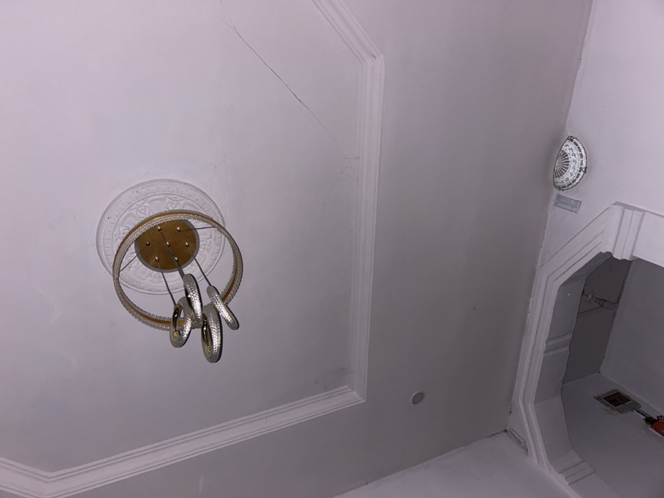 A low-angle shot reveals a white ceiling with decorative molding. A modern chandelier with gold accents and silver, swirling elements hangs from a circular medallion. A faint crack runs across the ceiling's surface. In the upper right, a white, decorative plate is mounted near a doorway with ornate molding and a glimpse inside reveals a dark interior space.